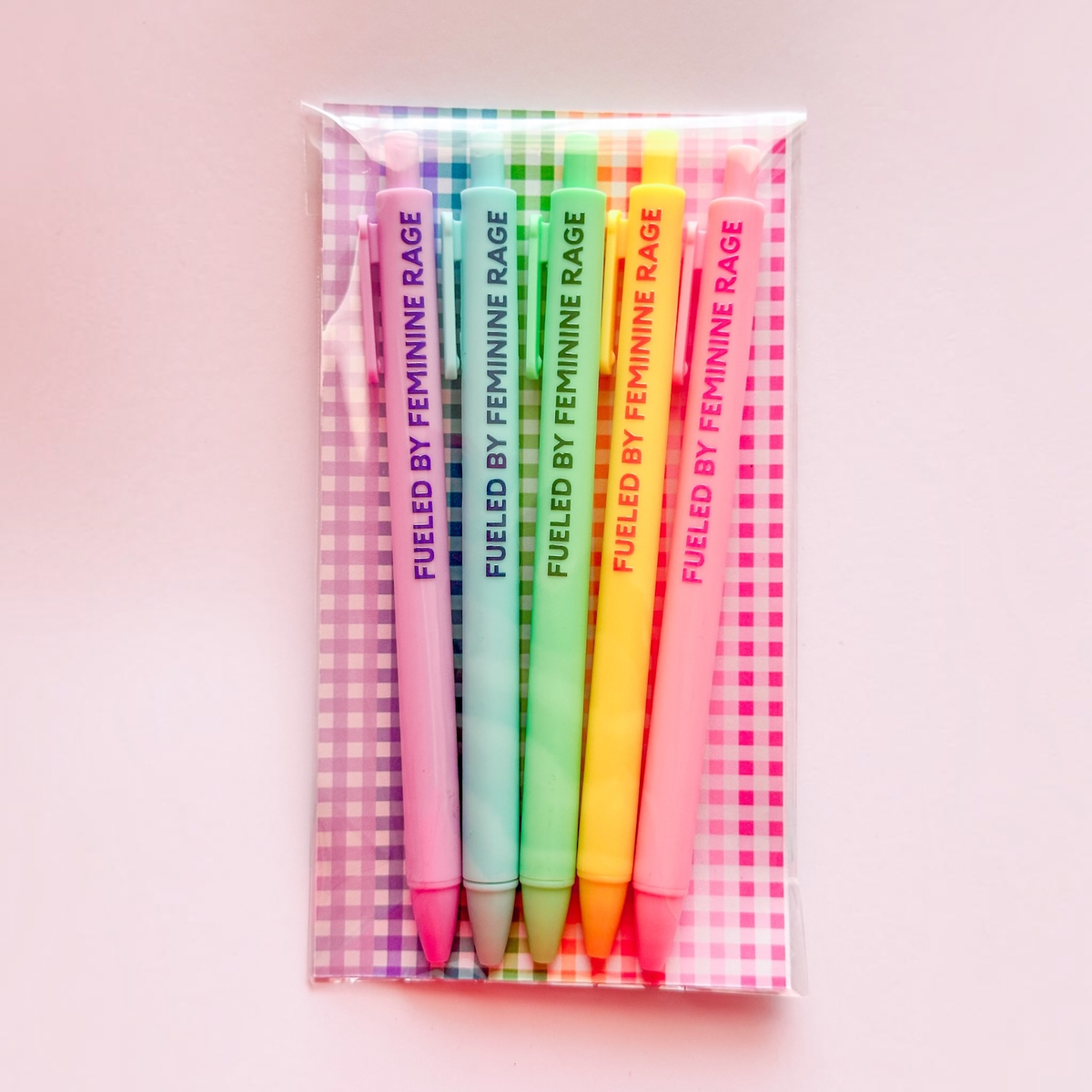A set of six colorful gel pens in a clear plastic sleeve with a pink and white gingham patterned backing. The pens are labeled 'FUELED BY FEMININE RAGE' in vertical text and come in shades of purple, blue, green, yellow, orange, and pink.