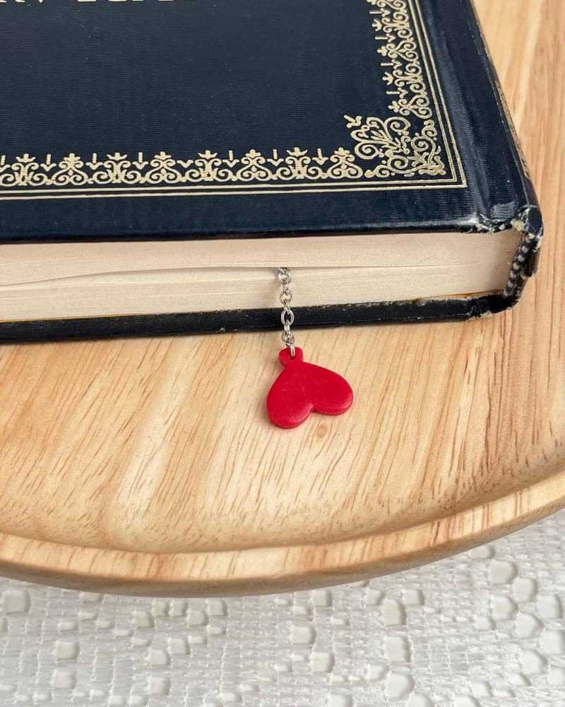 Red heart-shaped bookmark on a chain next to an open book on a wooden surface.