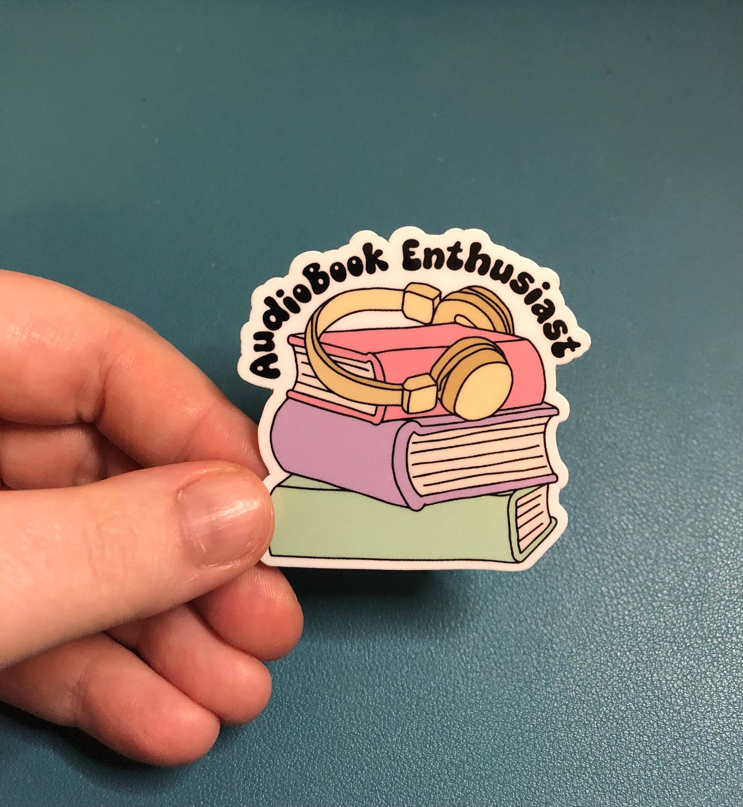 A hand holds a die-cut sticker featuring a stack of books with over-ear headphones on top and the words 'Audiobook Enthusiast' in a curved black outline. The sticker is shown against a textured teal background.
