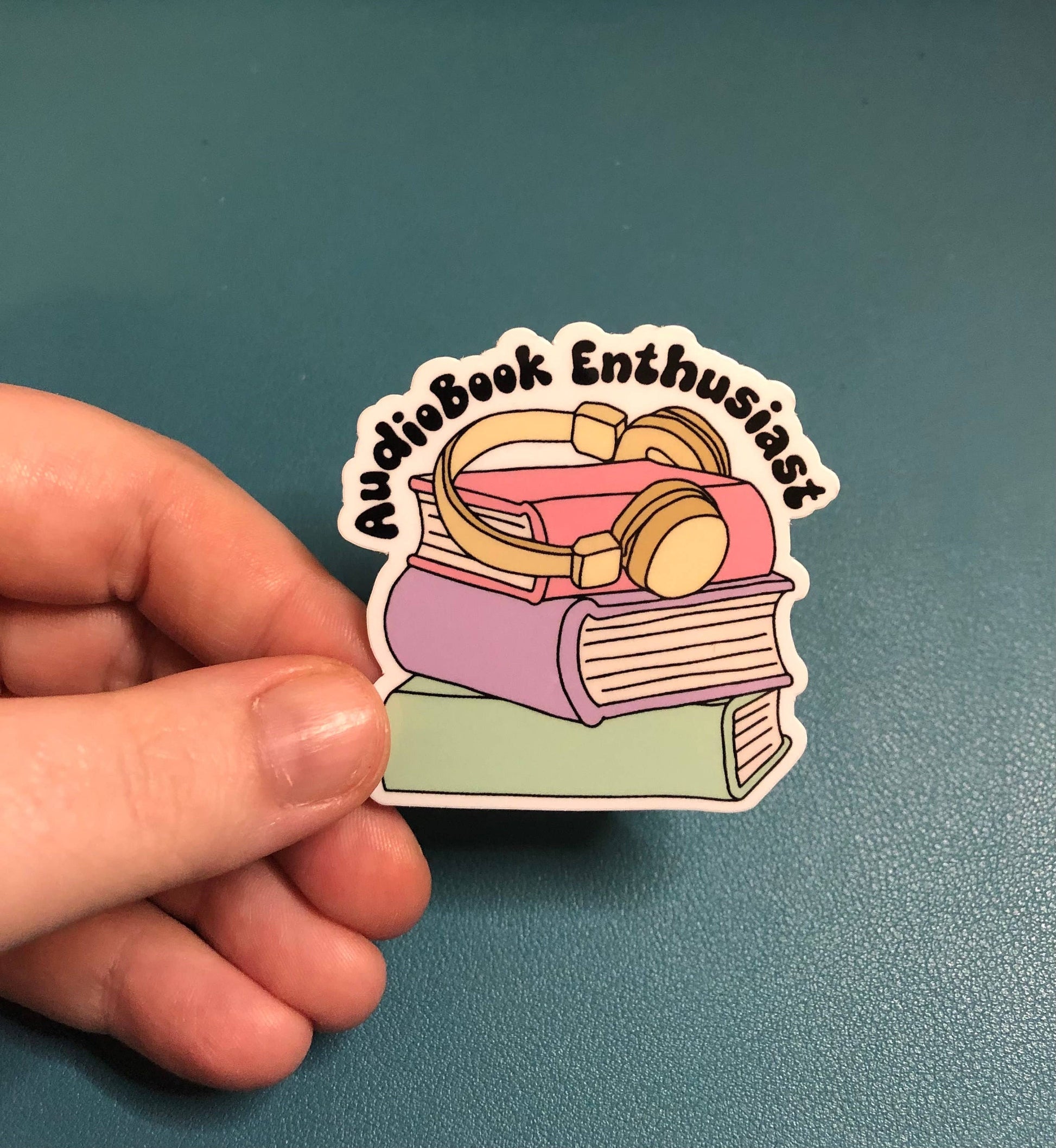 A hand holds a die-cut sticker featuring a stack of books with over-ear headphones on top and the words 'Audiobook Enthusiast' in a curved black outline. The sticker is shown against a textured teal background.