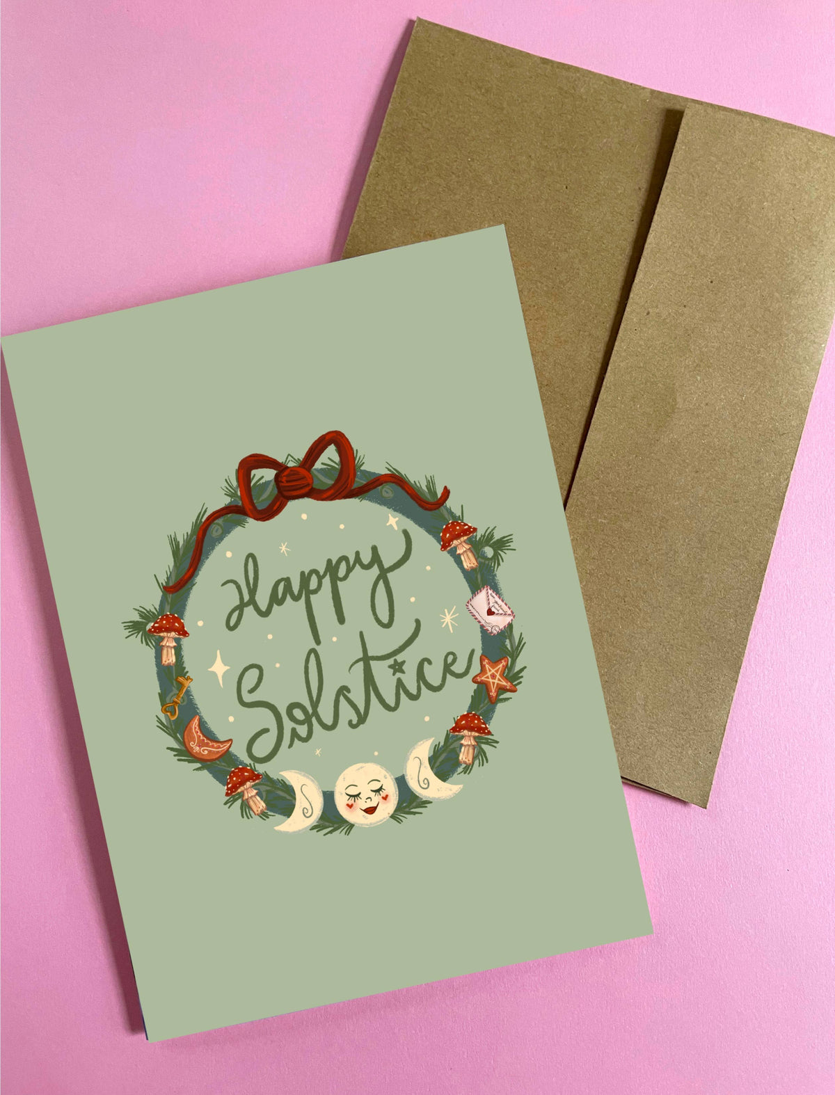 A square greeting card with a sage green background and a festive wreath illustration reading 'Happy Solstice' in script, shown with a brown kraft envelope on a pink background.