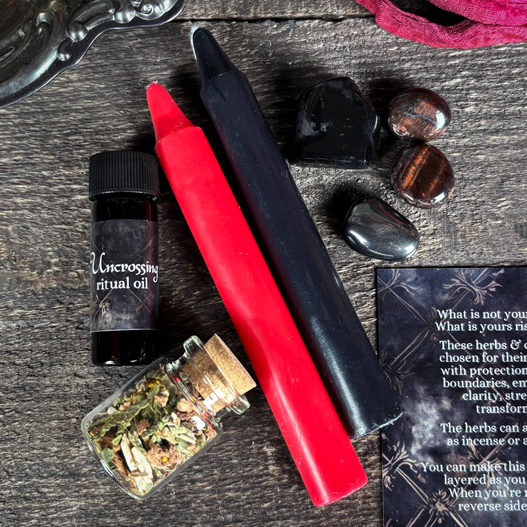 Red and black candles, herbs, and a bottle labeled 'Uncrossing ritual oil' on a wooden surface.