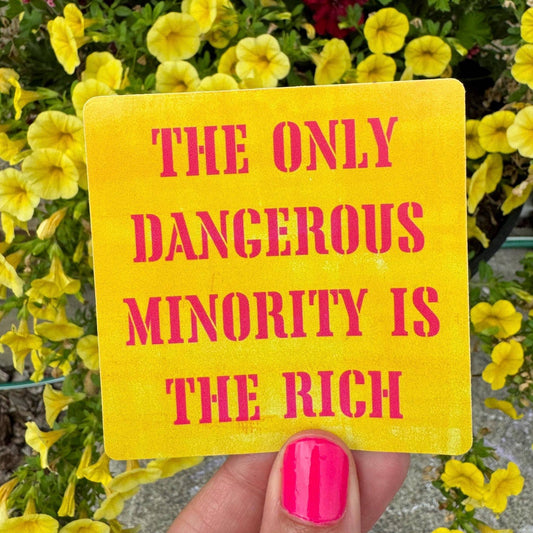 Yellow sign with pink text 'The Only Dangerous Minority is the Rich' held by a hand with pink nail polish, against a background of yellow flowers.