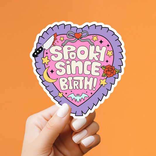 Heart-shaped sticker with 'Spooky Since Birth!' text held by a hand against an orange background