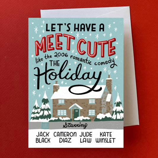 A personalized holiday greeting card with a light blue winter illustration, featuring the text 'LET'S HAVE A MEET CUTE like the 2006 romantic comedy THE Holiday' and a list of names at the bottom. The card is shown with a white envelope against a solid red background.