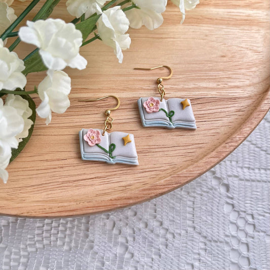 Book-shaped earrings with floral details on a wooden surface with white flowers.