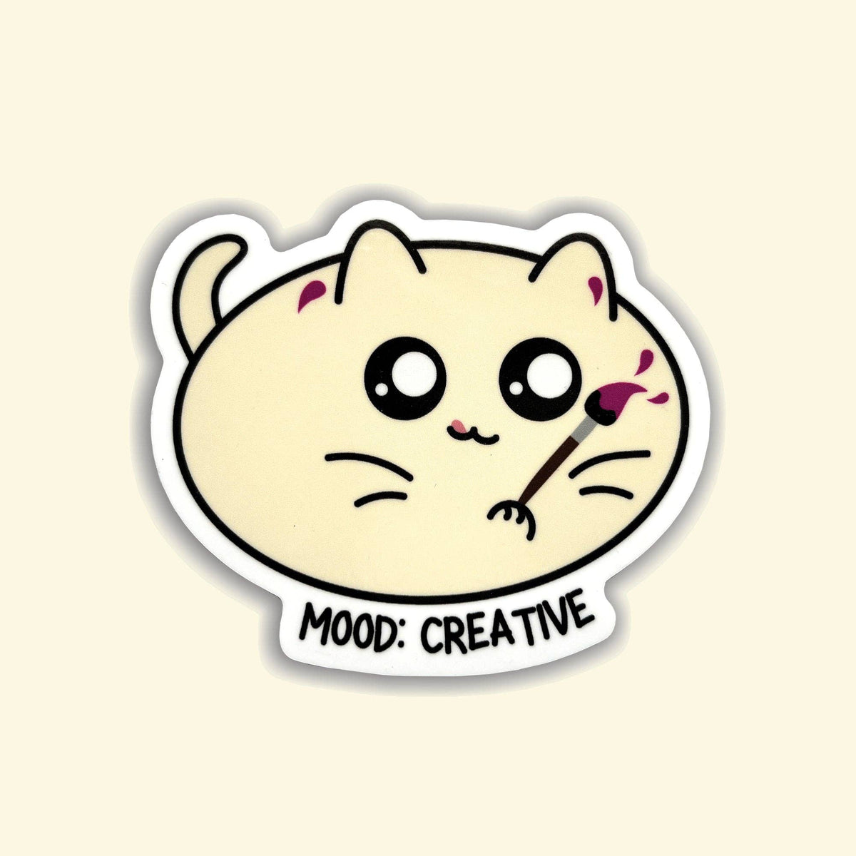 A die-cut sticker featuring a cute cream-colored cartoon cat with large black eyes, holding a paintbrush with pink paint, and the text 'MOOD: CREATIVE' below, shown on a light beige background.