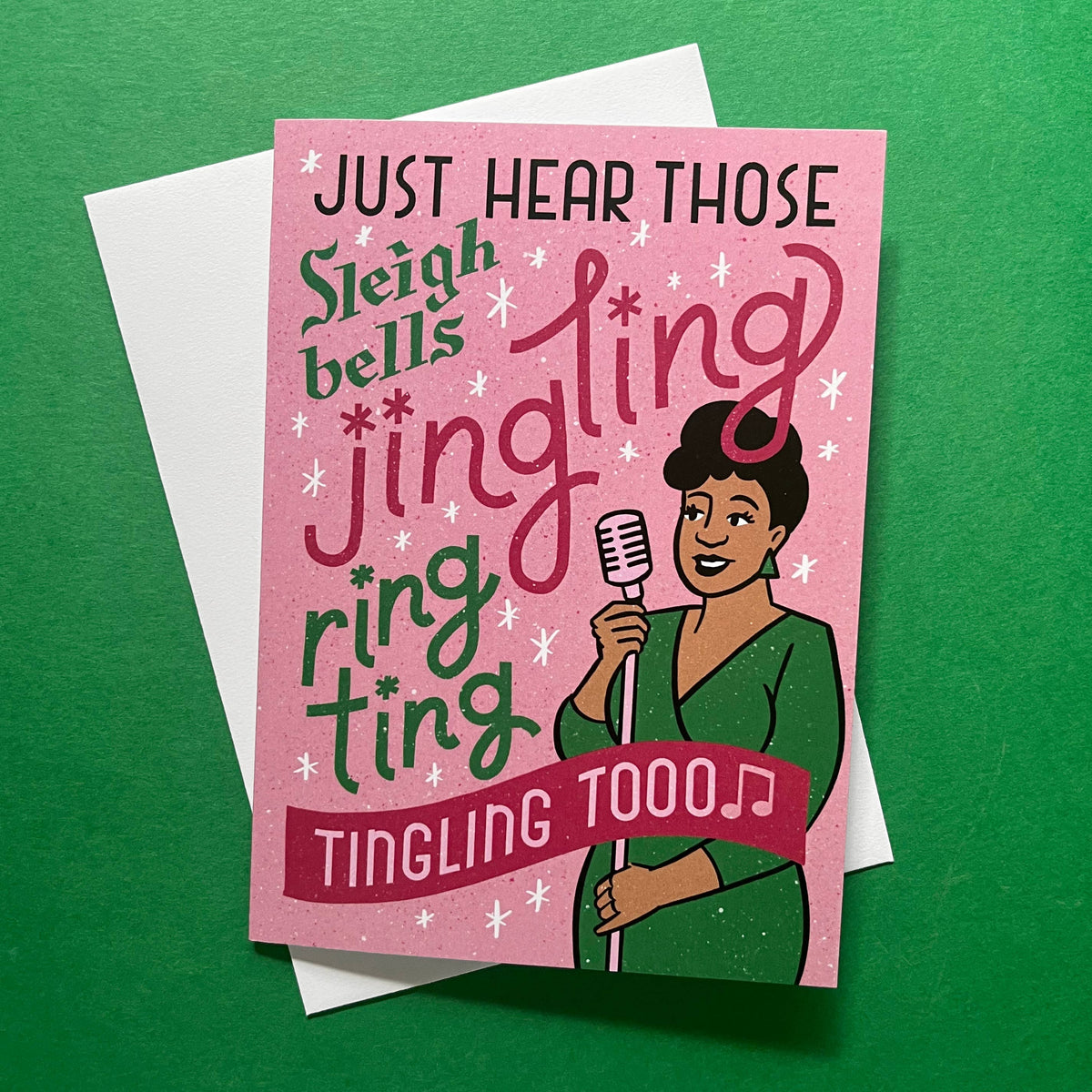 A pink greeting card with holiday-themed text reading 'JUST HEAR THOSE Sleigh bells jingling ring ting TINGLING TOOO' and an illustrated woman holding a microphone, shown with a white envelope on a green background.
