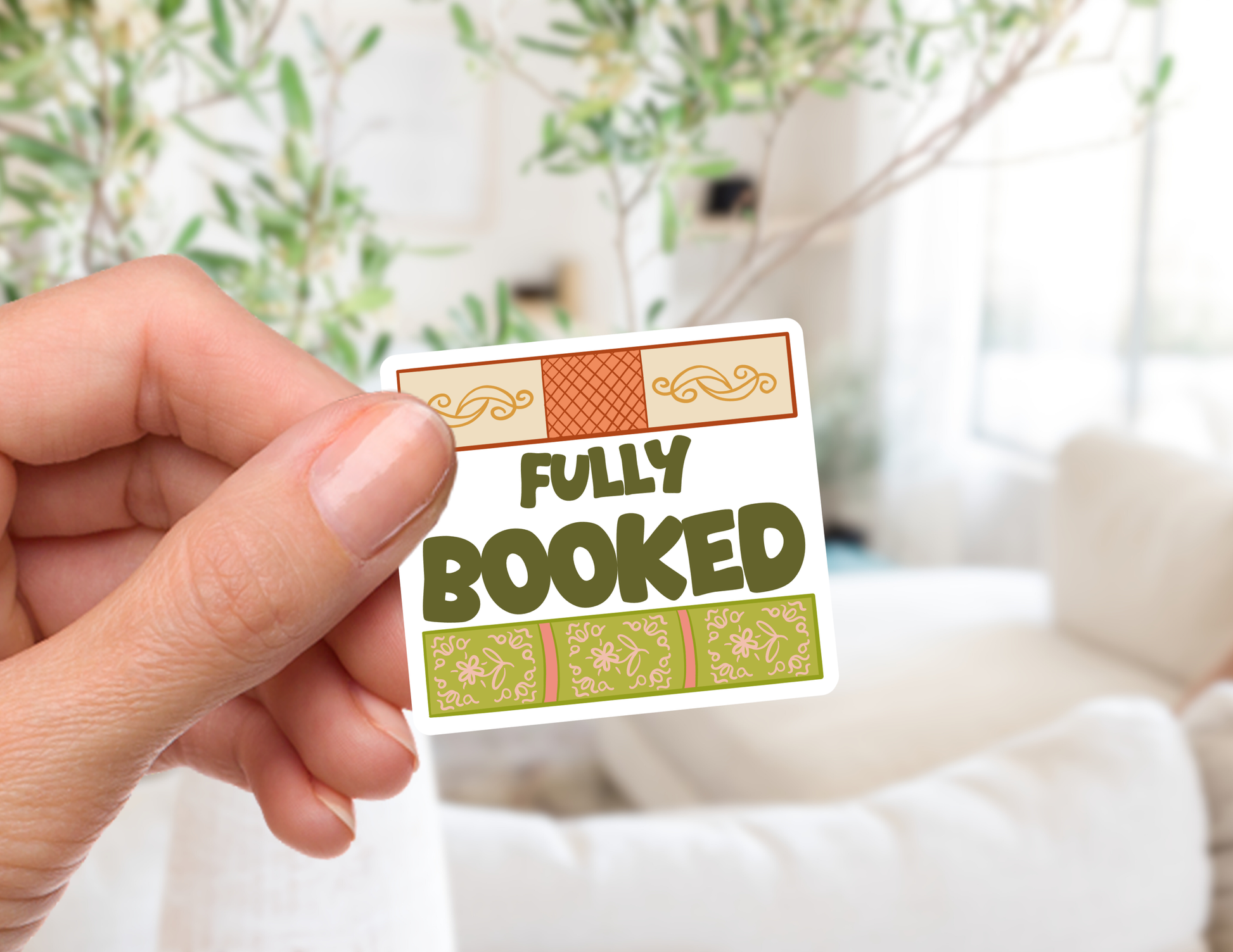 A hand holds a small rectangular sticker that reads 'FULLY BOOKED' in dark olive letters, with decorative beige and green patterned borders, against a softly blurred indoor background.
