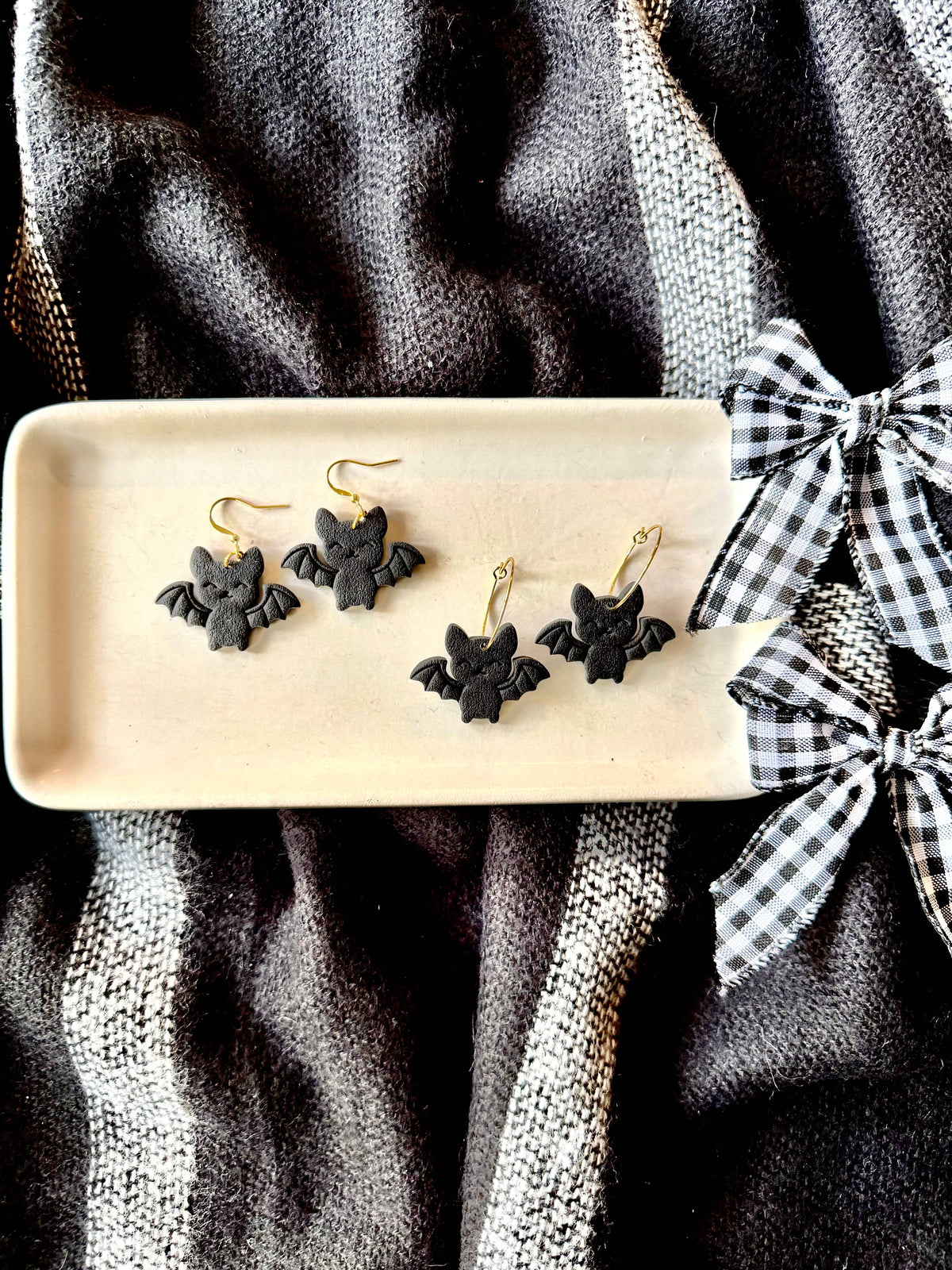 Three black bat-shaped dangle earrings with gold fishhook wires displayed on a beige ceramic tray, set against a dark textured fabric background with a black-and-white gingham ribbon bow.