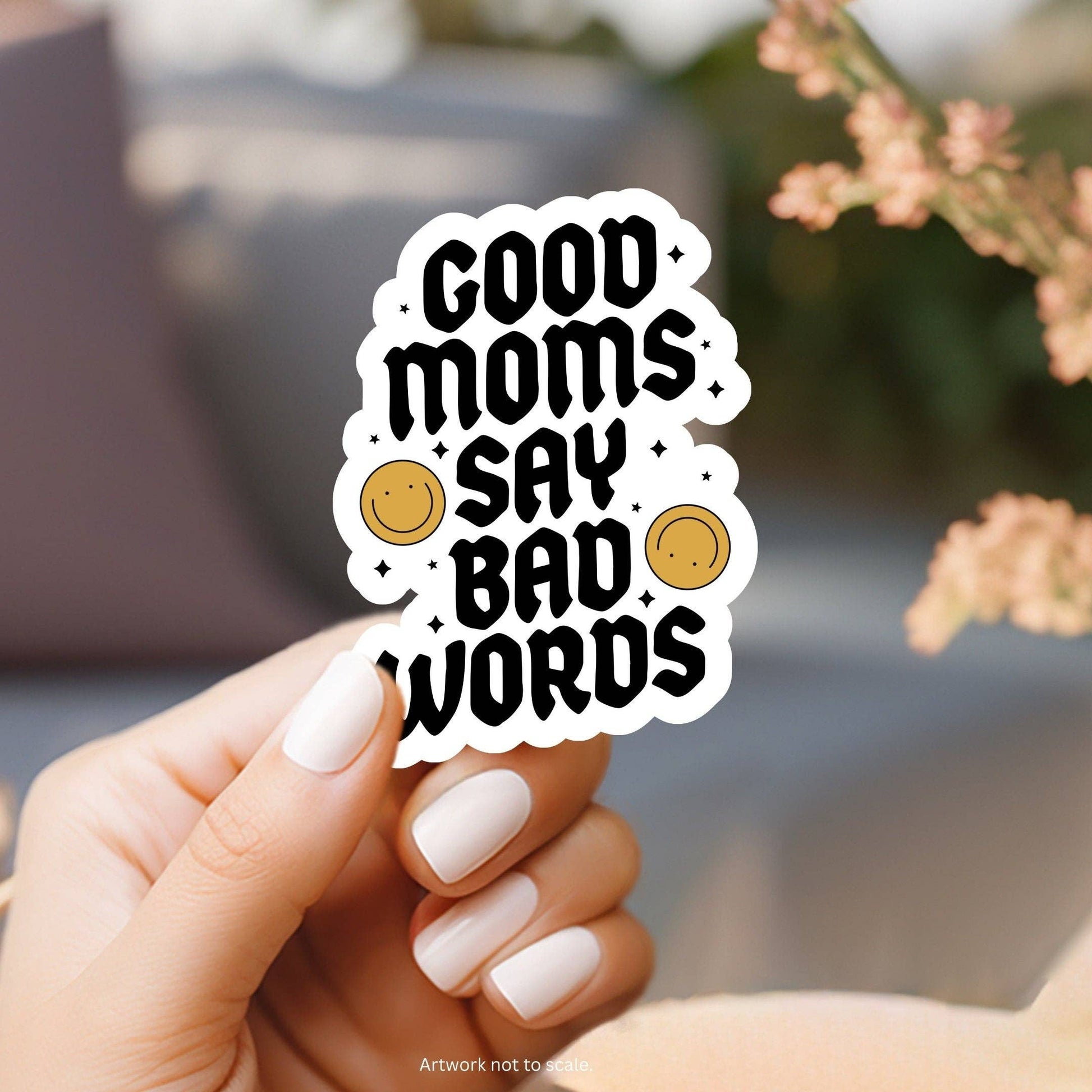 Hand holding a sticker with 'Good moms say bad words' text and smiley faces.