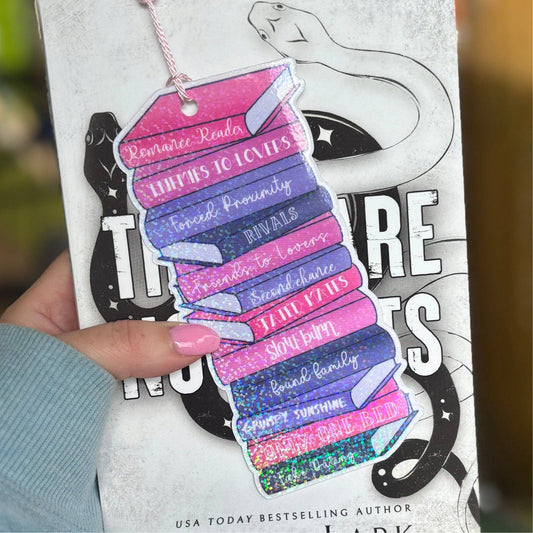 A cardstock bookmark with multiple sections, each printed with different text related to romance novel tropes, and a sparkle finish. The bookmark is held in front of a book.