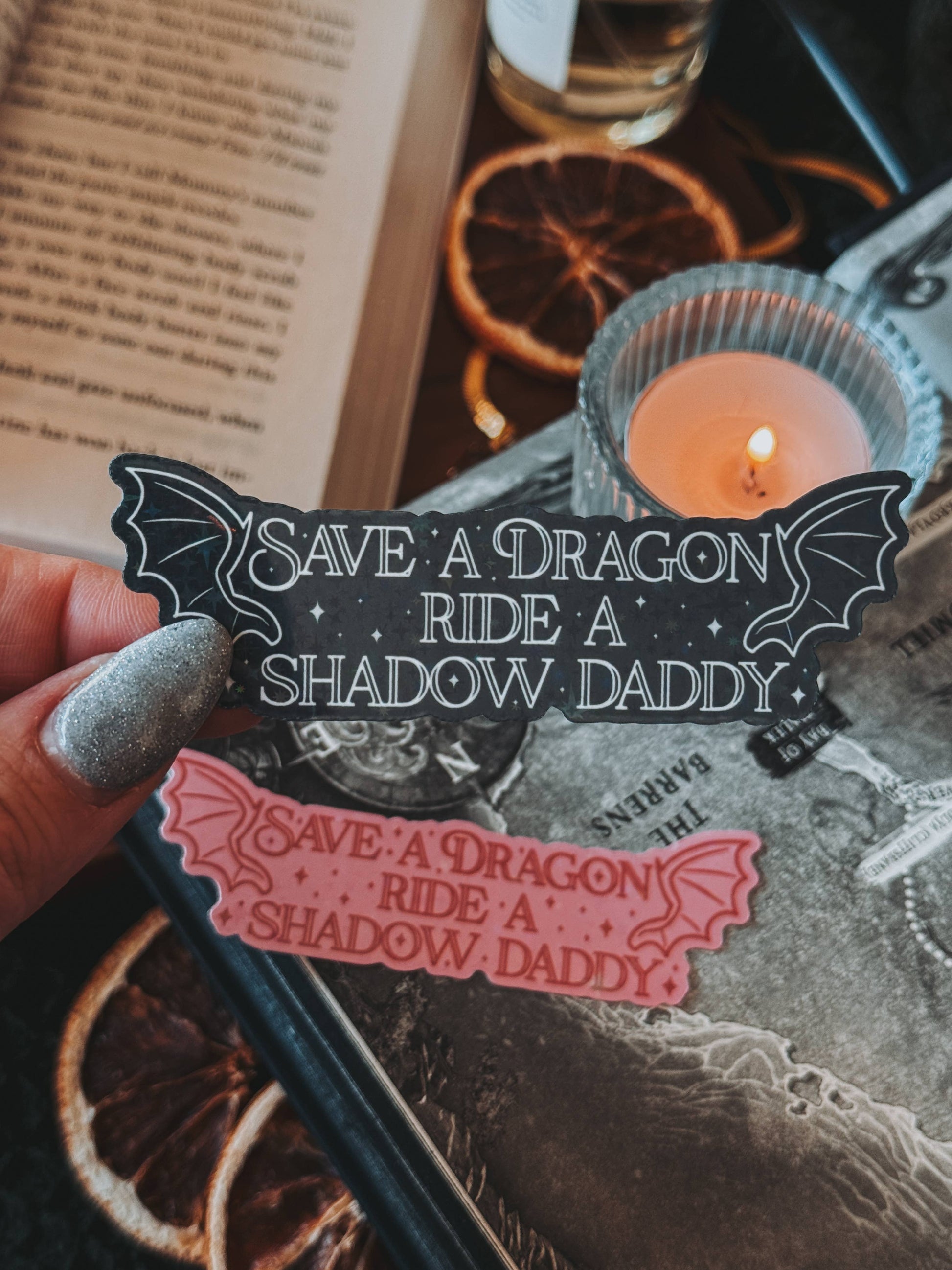 Sticker with 'Save a Dragon, Ride a Shadow Daddy' text held over a candlelit scene with an open book and dried oranges.