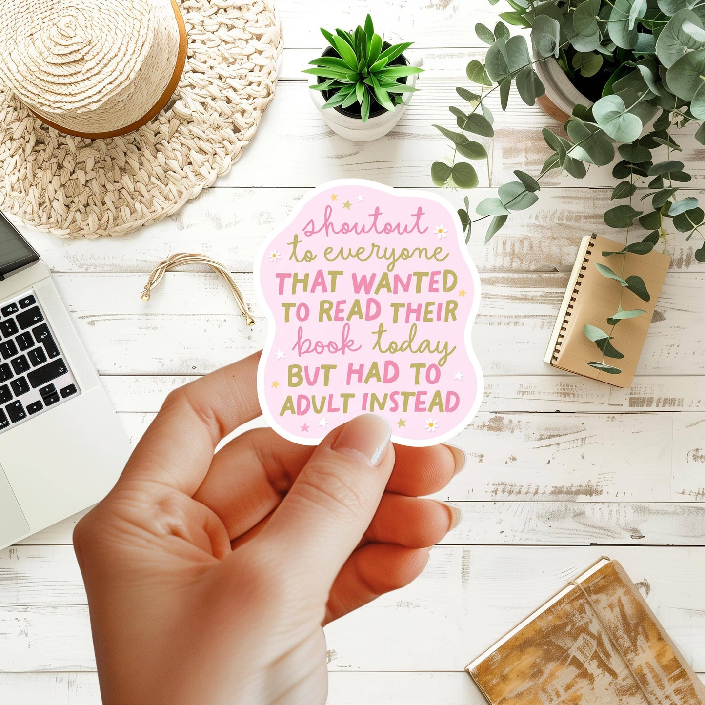 Hand holding a pink sticker with text on a desk with laptop, notebook, and plants
