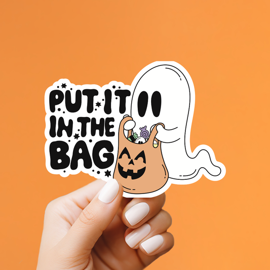 A hand with white manicured nails holds a die-cut sticker against a solid orange background. The sticker features the text 'PUT IT IN THE BAG' in black lettering next to a white ghost holding a jack-o'-lantern bag filled with candy.