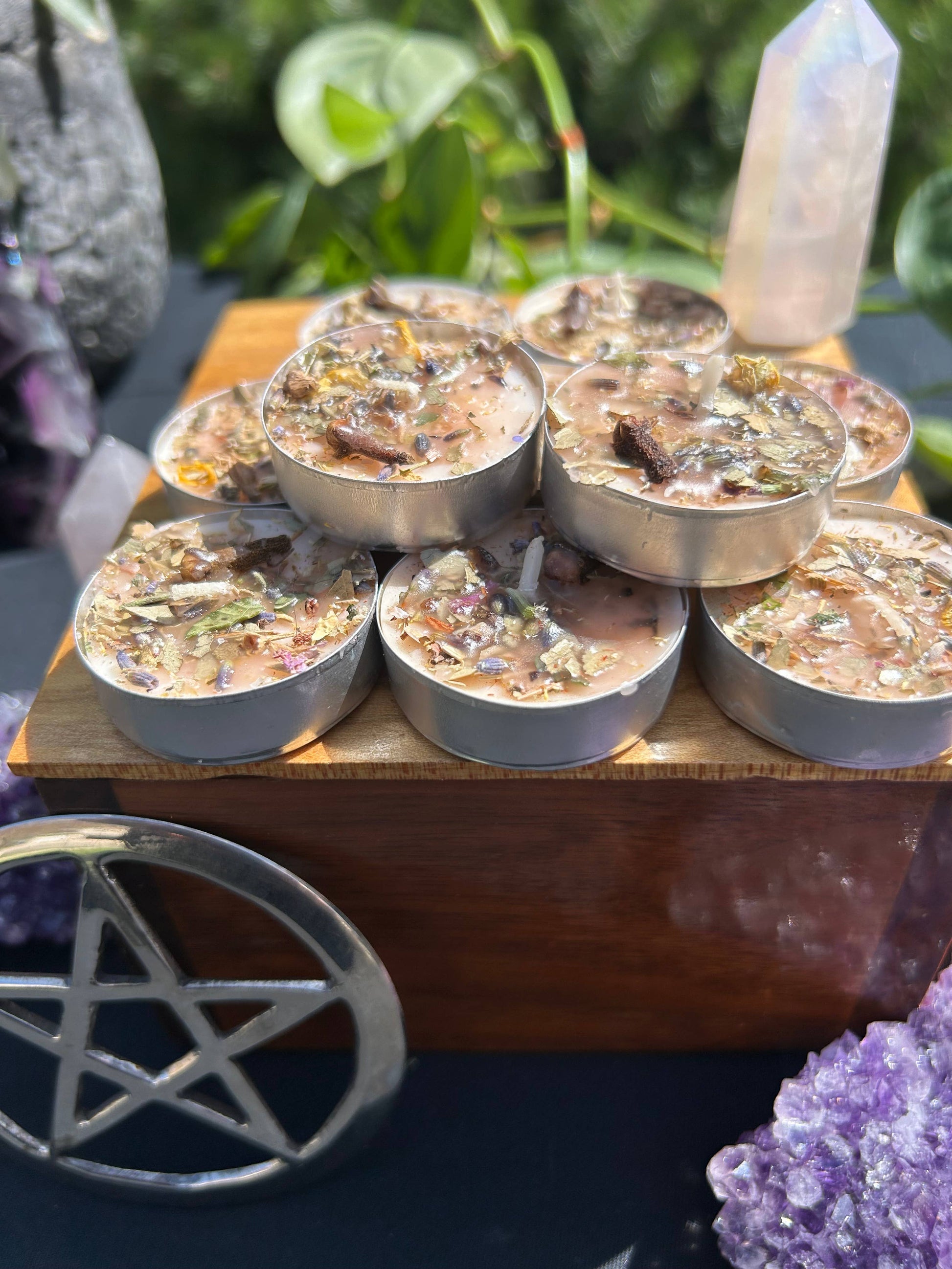 Set of small candles with herbs on a wooden stand, surrounded by crystals and plants.