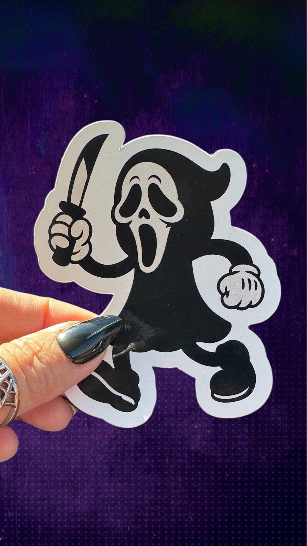 A die-cut vinyl sticker of the Ghostface character from the Ghostbusters franchise, held by a hand with black fingernails against a dark purple textured background.
