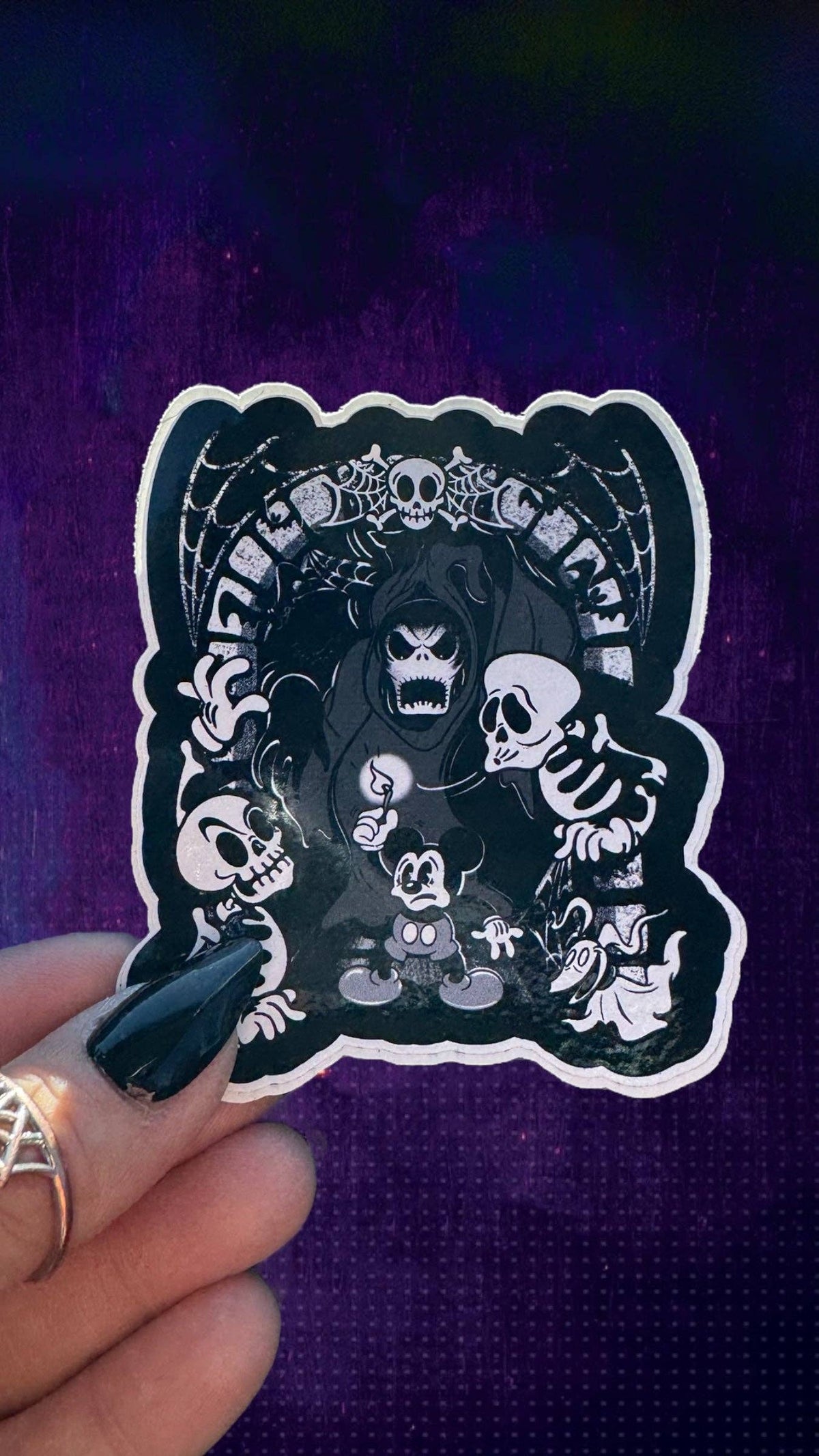 A hand with black nail polish holds a die-cut sticker featuring a black and white gothic cartoon illustration of Mickey Mouse surrounded by skulls, skeletons, and a grim reaper, set against a dark purple textured background.