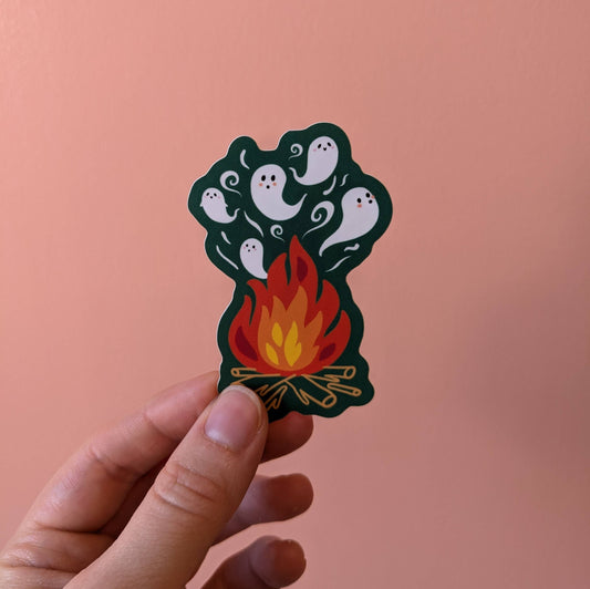 A hand holding a die-cut sticker featuring a dark green silhouette with white cartoon ghosts rising above a red, orange, and yellow campfire, set against a solid peach-pink background.