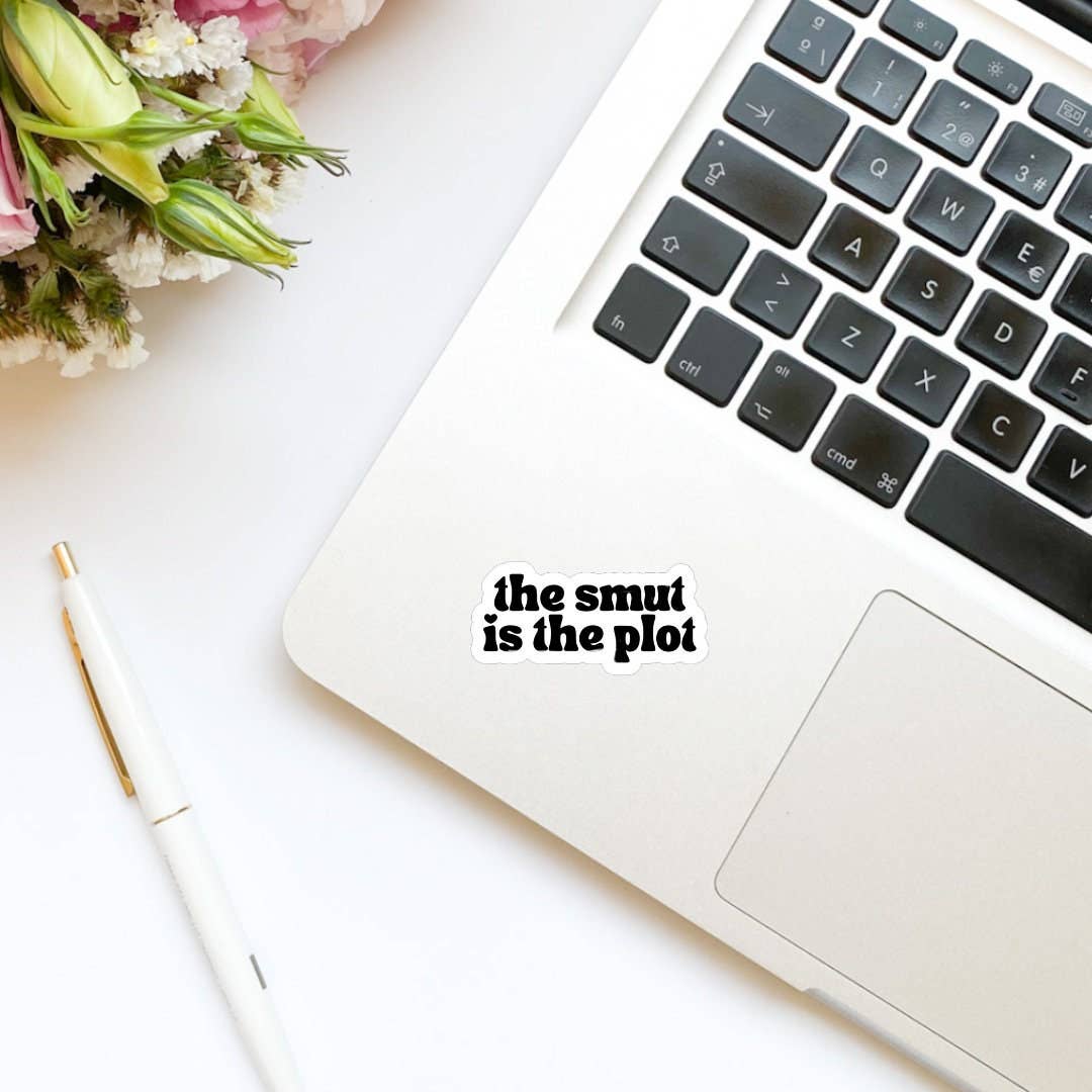 Laptop with 'the smut is the plot' sticker on a white surface with flowers and a pen.
