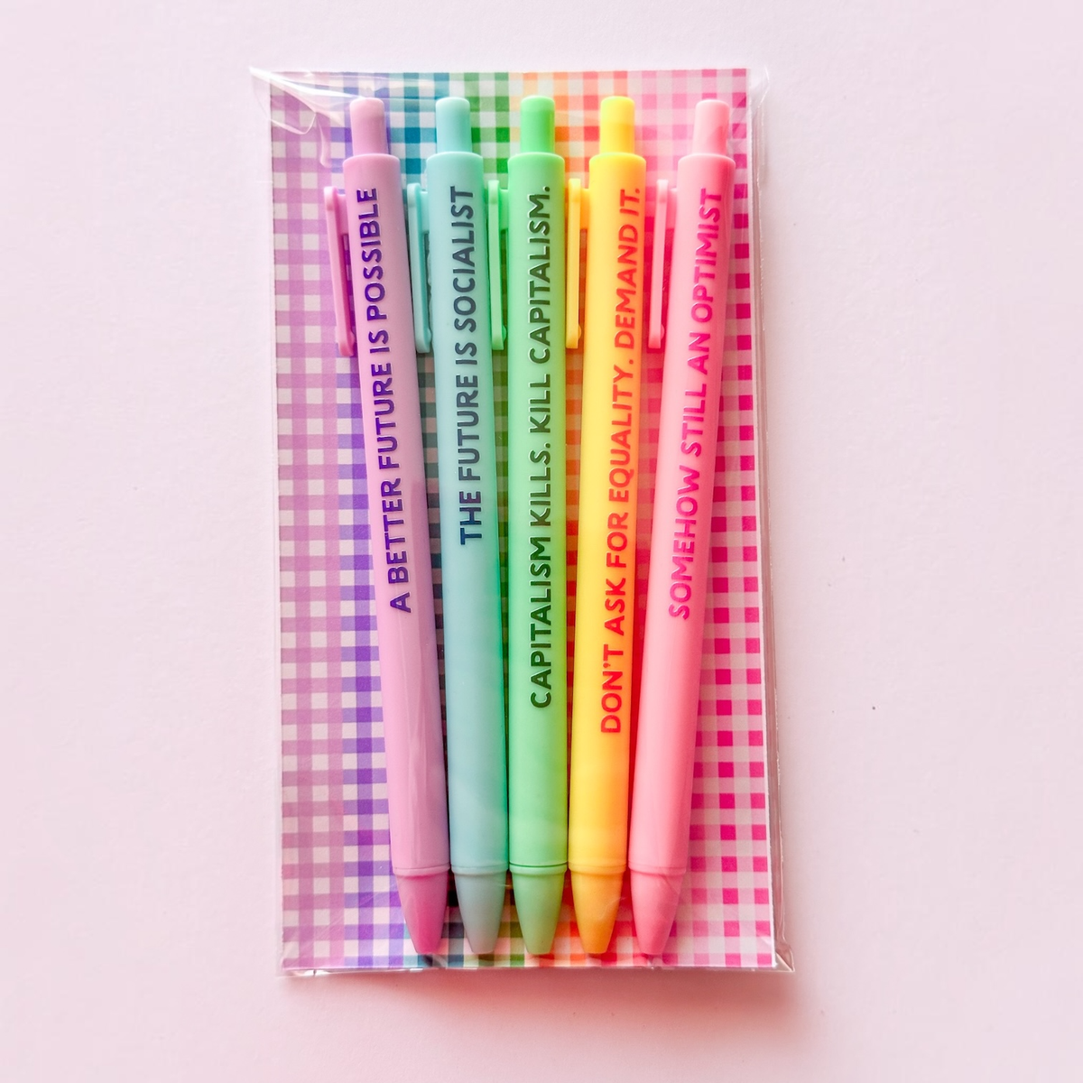 A set of five pastel-colored pens in a clear plastic sleeve with a pink and green gingham patterned backing card. The pens are pink, blue, green, yellow, and orange, each printed with a political slogan in vertical text.