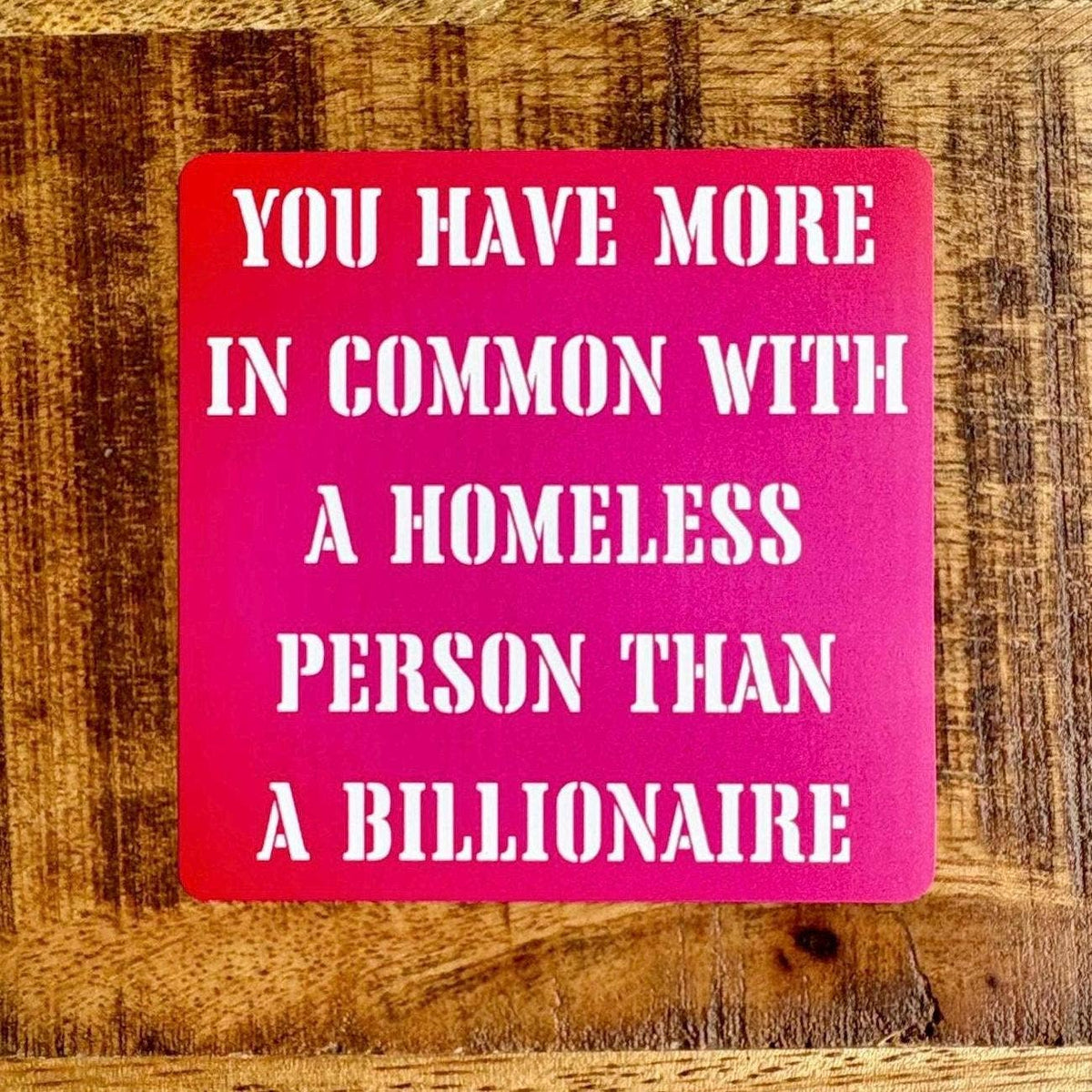A square pink sign with bold white text reading 'YOU HAVE MORE IN COMMON WITH A HOMELESS PERSON THAN A BILLIONAIRE' placed on a rustic wooden surface.