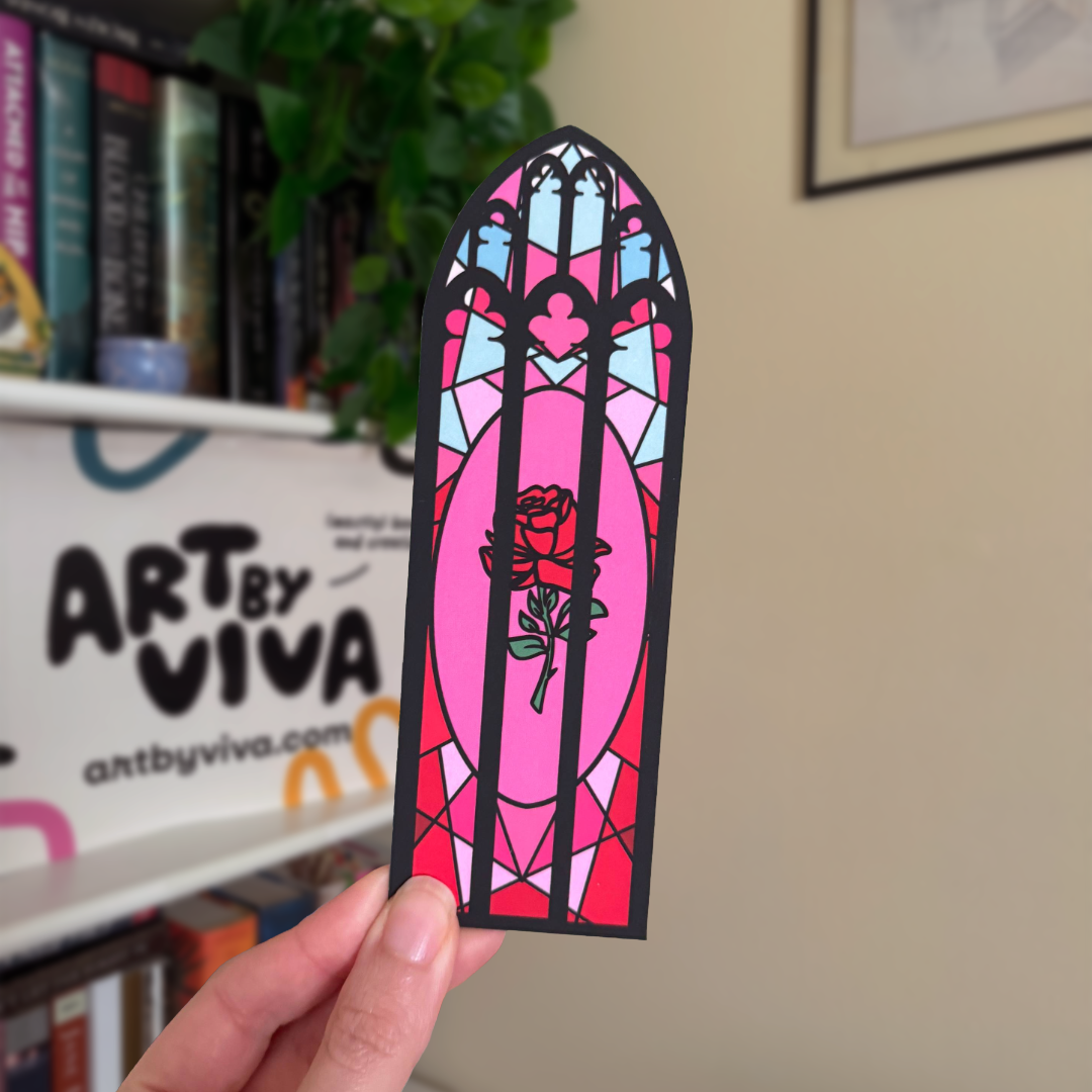 Stained glass-style bookmark with a rose design held by a hand in front of a bookshelf with 'Art by Viva' branding.
