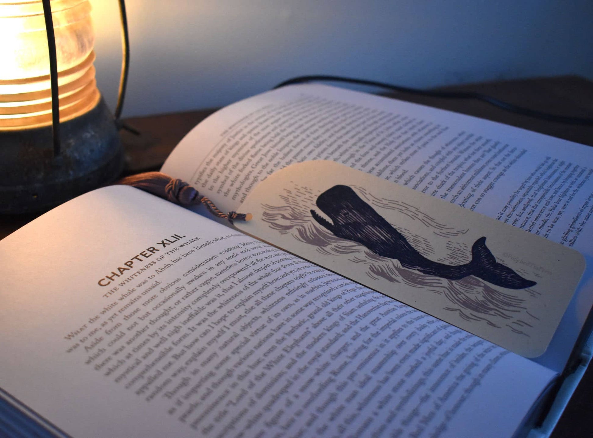 Open book with a whale bookmark on a wooden surface next to a lamp.