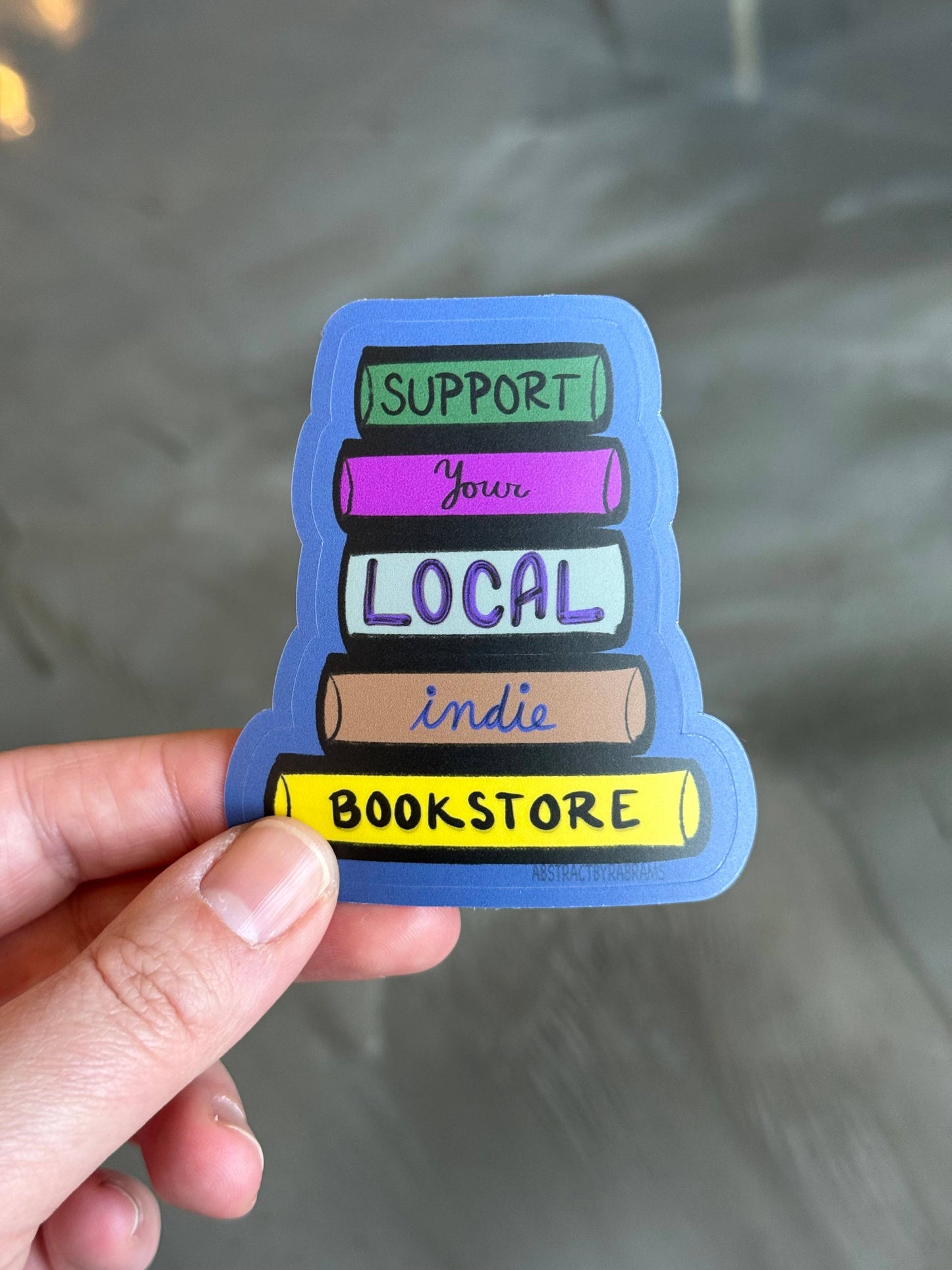 Sticker with 'Support Your Local Indie Bookstore' text held by a hand against a reflective surface.