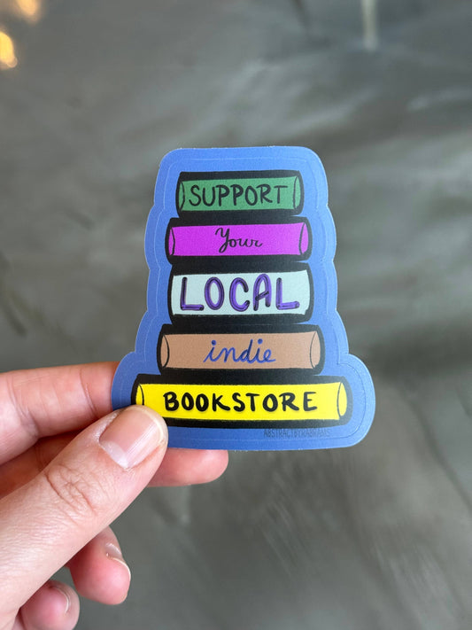 Sticker with 'Support Your Local Indie Bookstore' text held by a hand against a reflective surface.