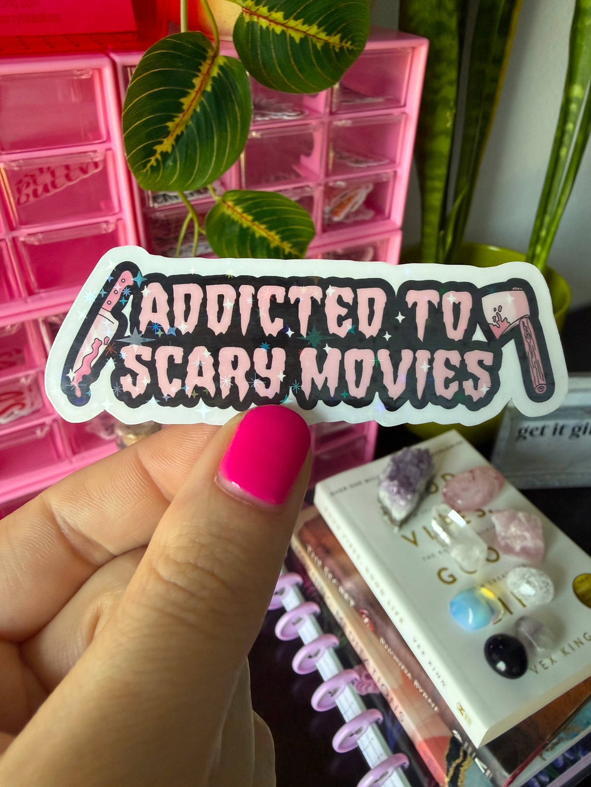 A hand with bright pink nail polish holds a die-cut sticker that reads 'ADDICTED TO SCARY MOVIES' in stylized pink and black lettering, with small horror-themed illustrations on both sides. The background is a cozy desk setup with pink storage drawers, green plants, a notebook, and crystals.