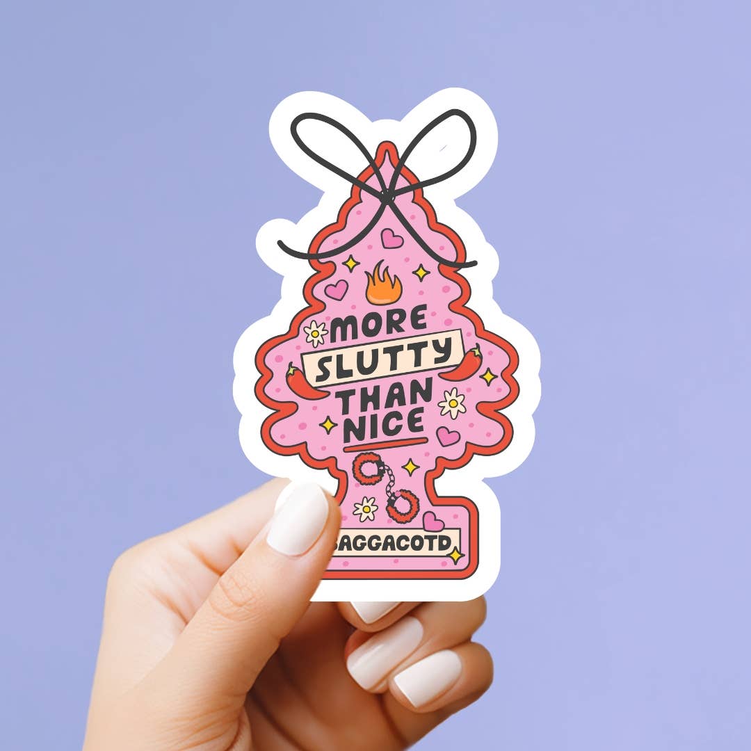 A hand with white manicured nails holds a die-cut sticker shaped like a pink gift box with a bow. The sticker features the text 'MORE SLUTTY THAN NICE' in black letters, decorated with small hearts, sparkles, and a small tag at the bottom that reads 'GAGCACOTD.