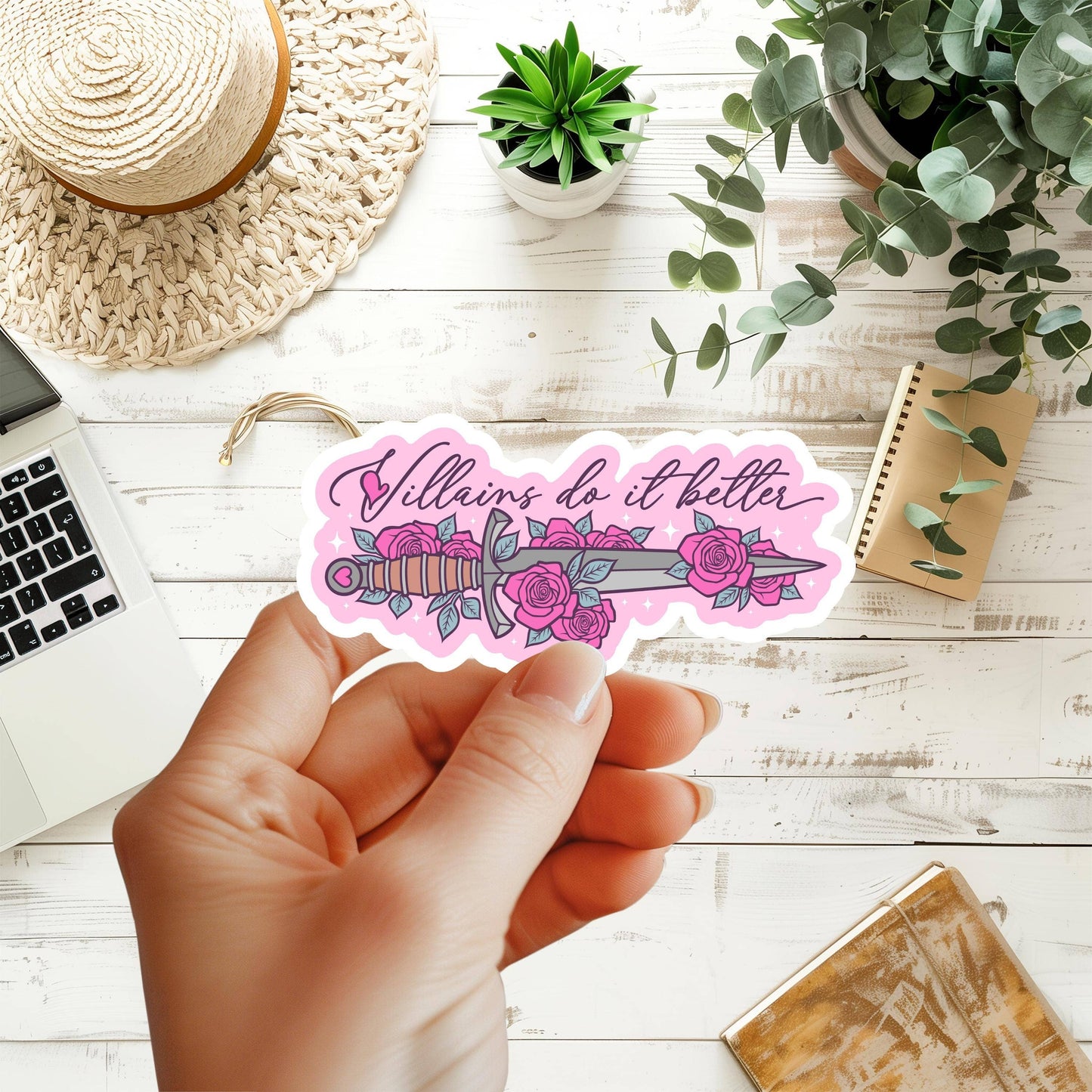 A hand holds a die-cut sticker featuring a pink and purple dagger surrounded by roses with the phrase 'Villains do it better' in script. The scene is styled on a white wood tabletop with a laptop corner, woven hat, small succulent, trailing greenery, a notebook, and a gold-toned box.