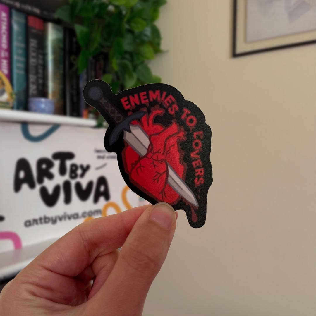 Hand holding a sticker with 'Enemies to Lovers' design in front of a shelf with books and a sign reading 'Art by Viva'.