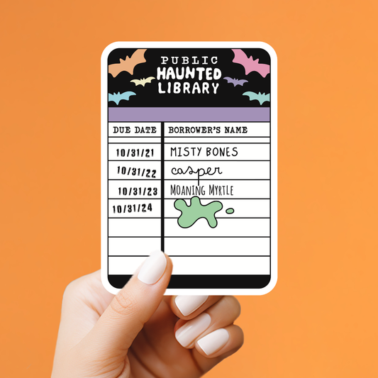 A hand with white manicured nails holds a rectangular sticker against a solid orange background. The sticker is designed like a 'Public Haunted Library' due date list with black, purple, and white sections, decorated with bats and a green slime graphic.