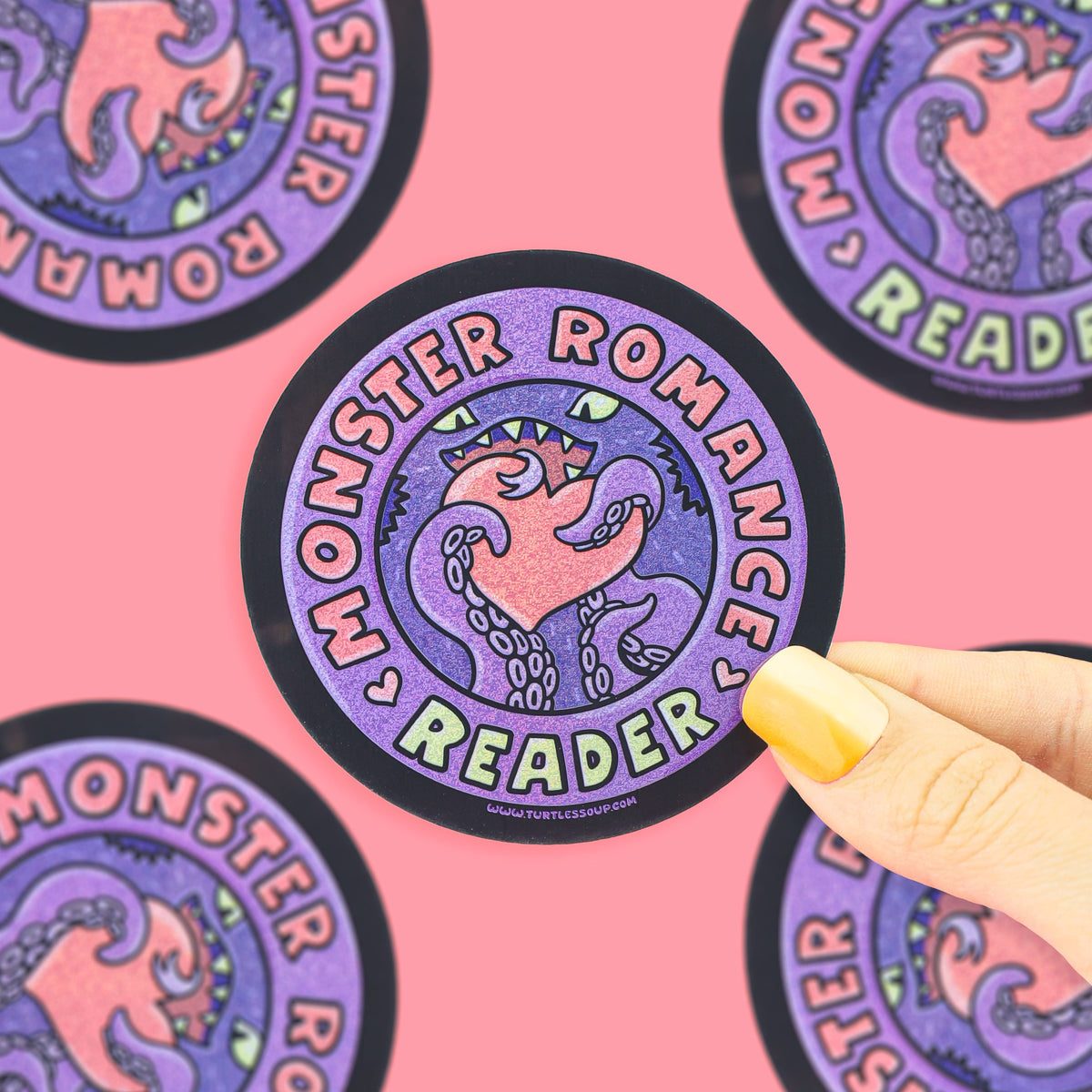 A circular sticker with a purple and pink monster illustration and the words 'Monster Romance Reader' is held by a hand with yellow nail polish against a pink background with other stickers visible.