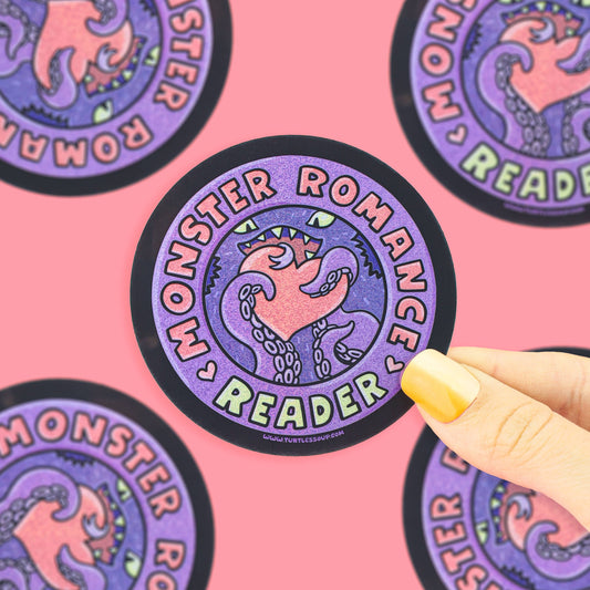 A circular sticker with a purple and pink monster illustration and the words 'Monster Romance Reader' is held by a hand with yellow nail polish against a pink background with other stickers visible.