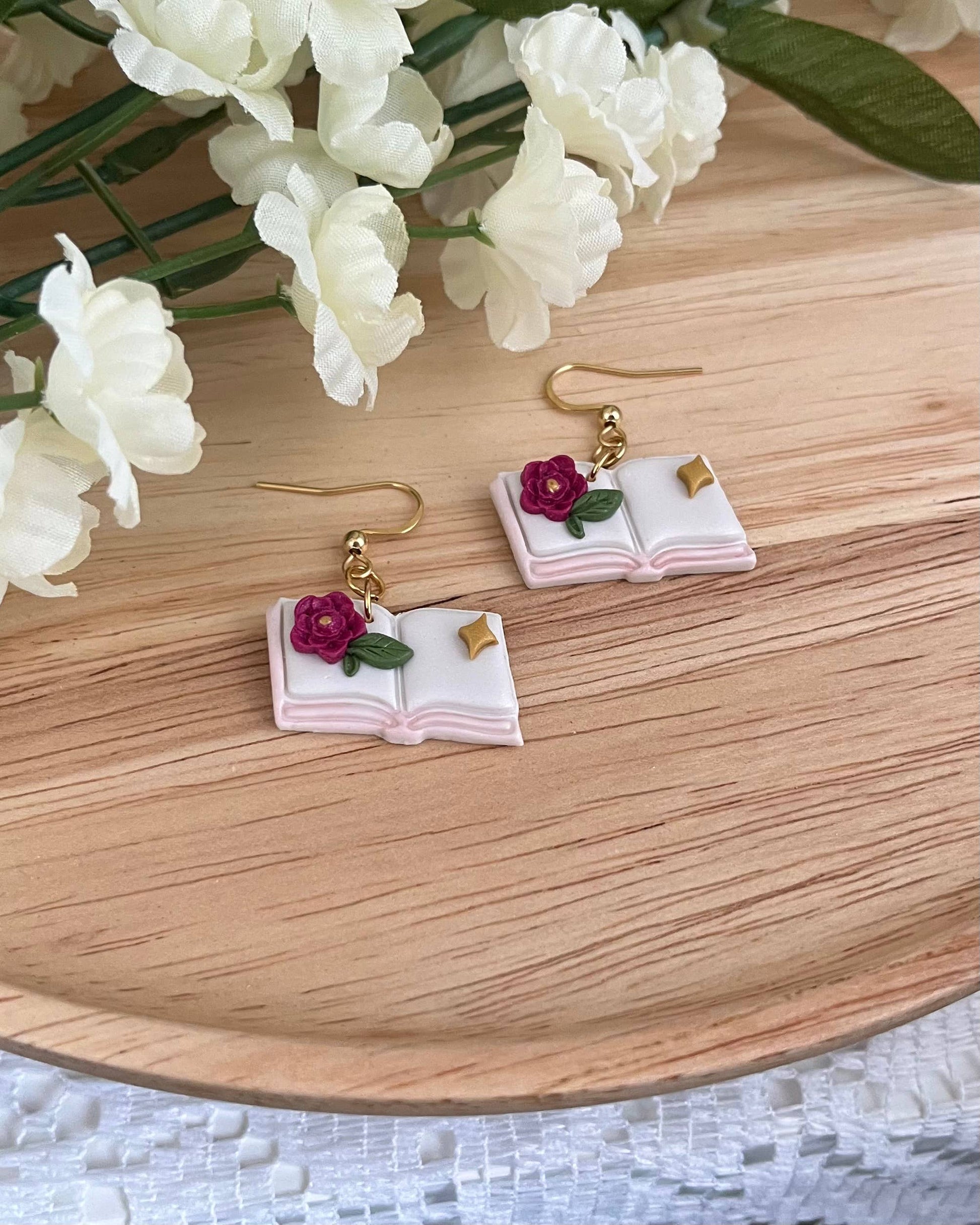 Book-shaped earrings with floral details on a wooden surface with white flowers.