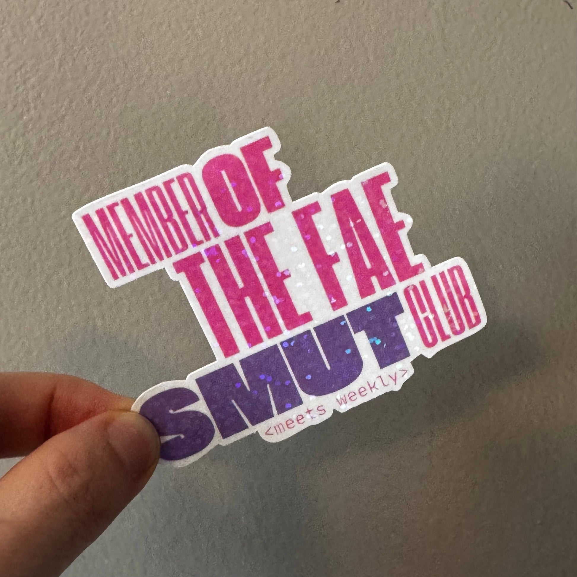 A hand holding a die-cut sticker that reads 'MEMBER OF THE FAE SMUT CLUB' with small text '·meets weekly' underneath, shown against a plain gray wall.
