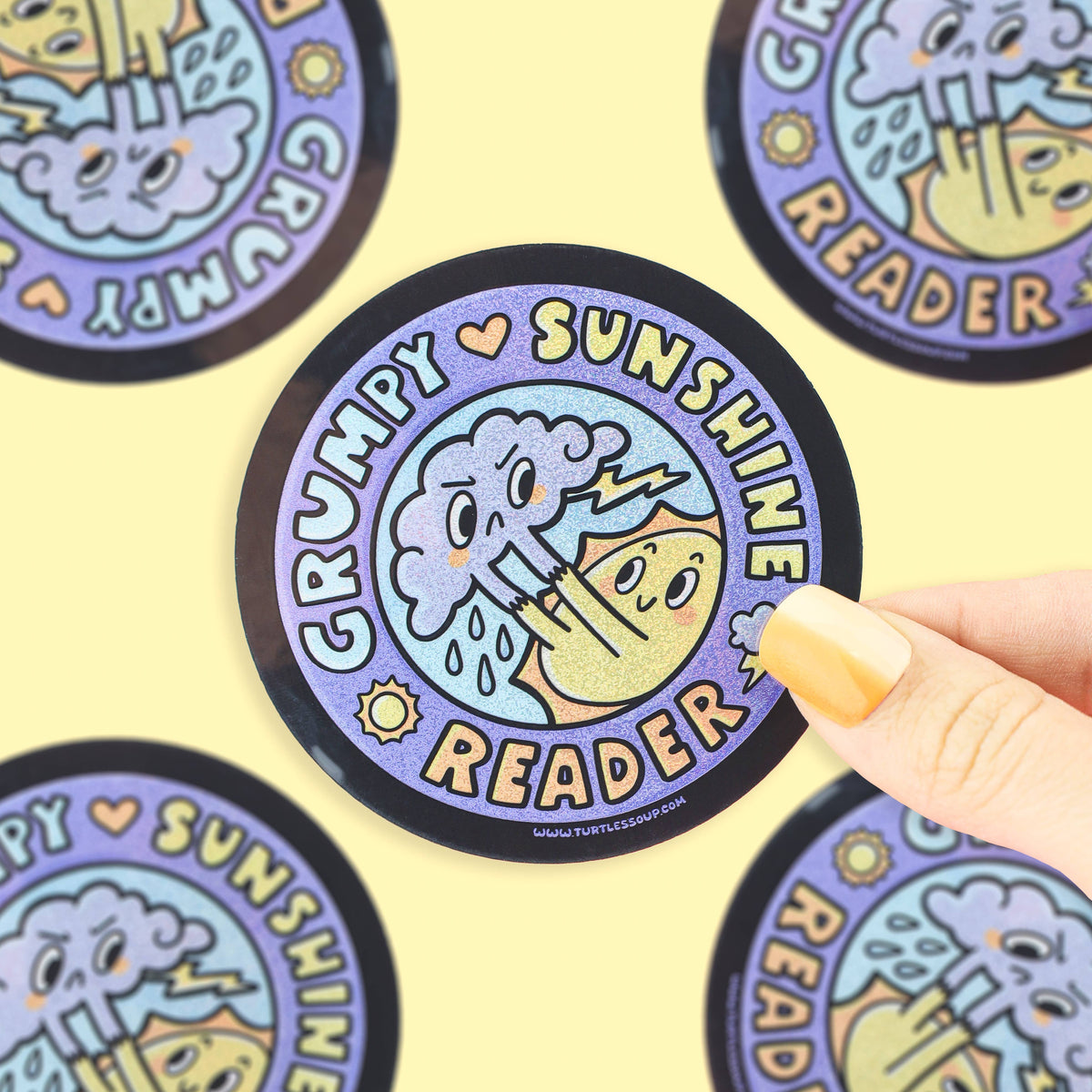 A circular sticker or coaster featuring a cartoon grumpy sunshine reader character, held by a hand with yellow nail polish against a pale yellow background with other copies of the sticker visible.