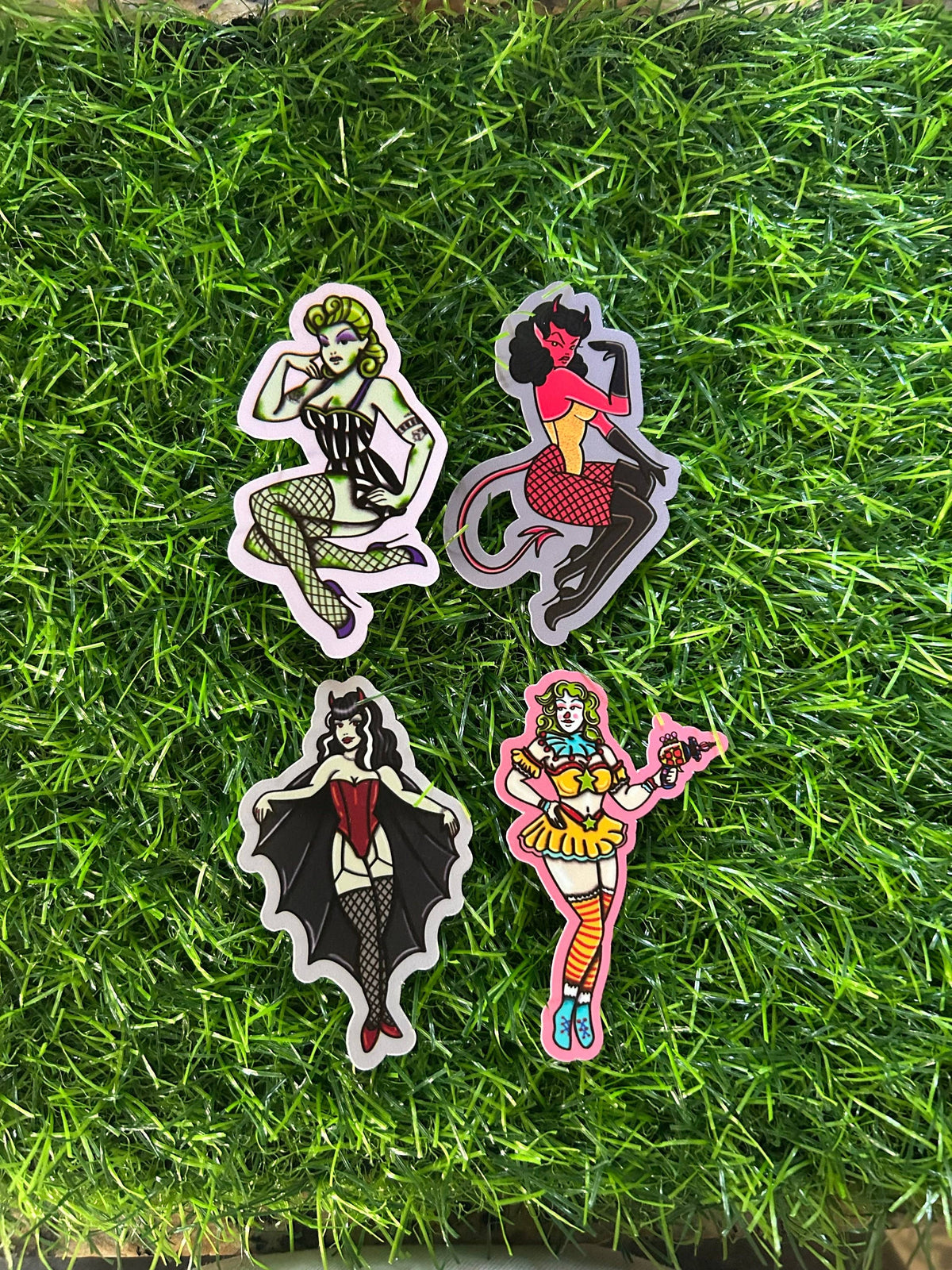Four die-cut vinyl stickers featuring stylized gothic and devil-themed female characters, arranged on a green grass background.