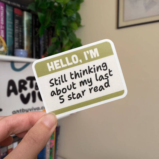 Hand holding a sticker with text 'HELLO, I'M Still thinking about my last 5 star read' in front of a bookshelf.