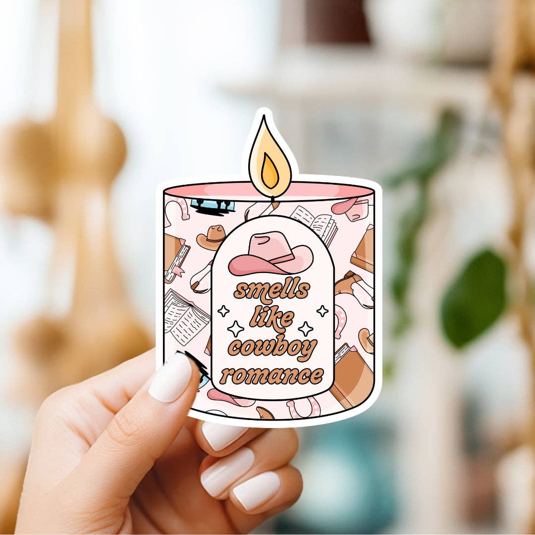 Sticker of a candle labeled 'smells like cowboy romance' held by a hand.