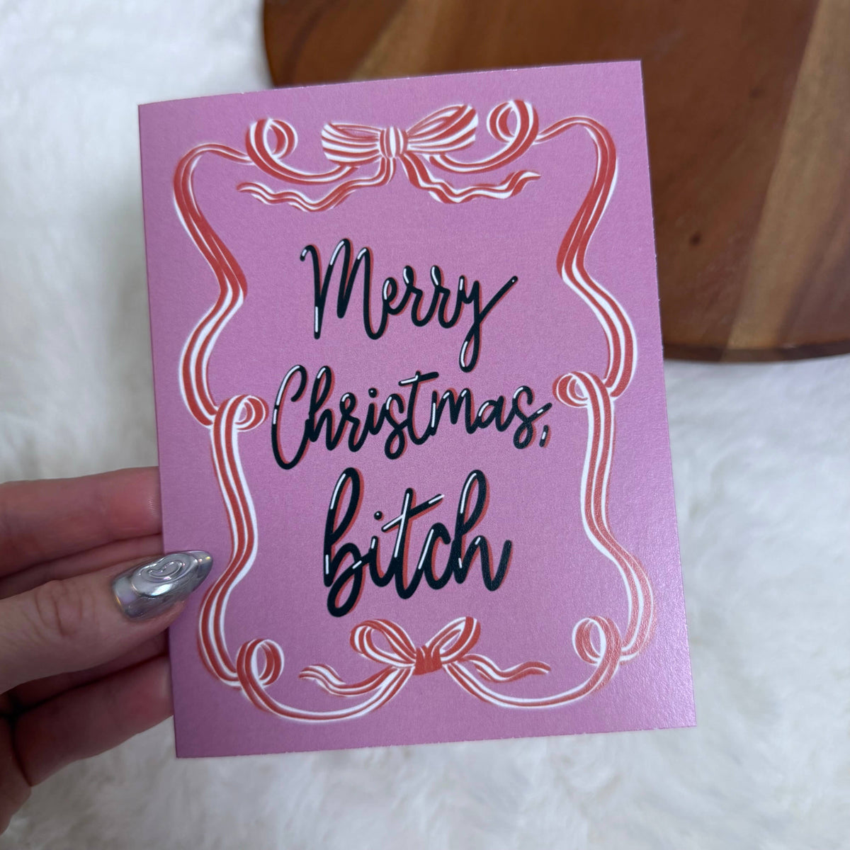A hand holding a pink Christmas greeting card with metallic rose-gold foil scrollwork and black cursive text reading 'Merry Christmas, bitch' on a white faux-fur surface.