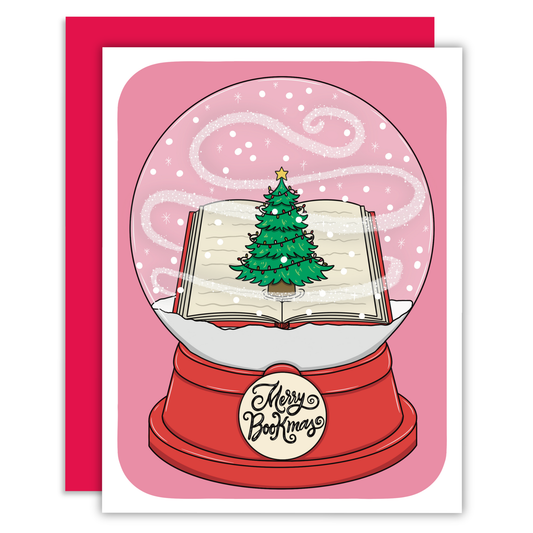 A Christmas greeting card featuring an illustration of an open book inside a snow globe with a green Christmas tree and falling white snow, with a red base that reads 'Merry, Merry Christmas.' A red envelope is partially visible behind the card.