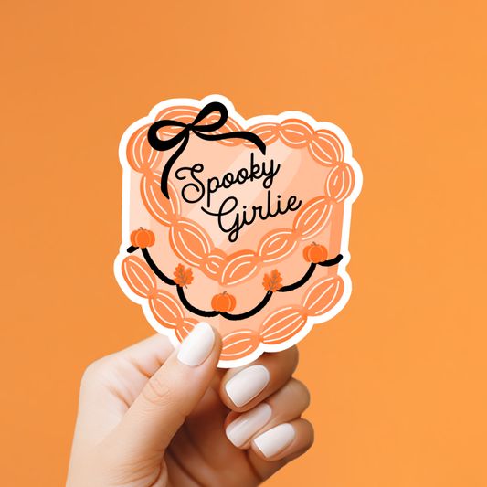 A hand with white manicured nails holds a scalloped die-cut sticker that reads 'Spooky Girlie' in black script, featuring an orange pumpkin wreath design with black outlines, against a solid orange background.