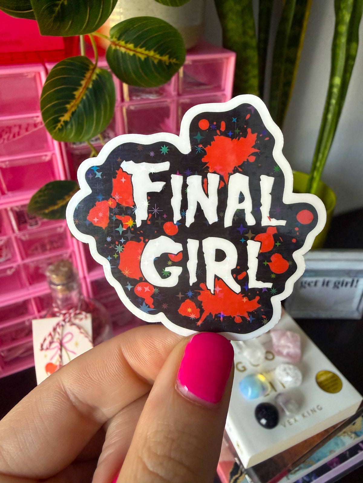A hand with bright pink nail polish holds a die-cut sticker that reads 'FINAL GIRL' in white letters over a black background with red paint splatters and small star details. The background is a colorful, cluttered desk with pink drawers, green plants, and other items.