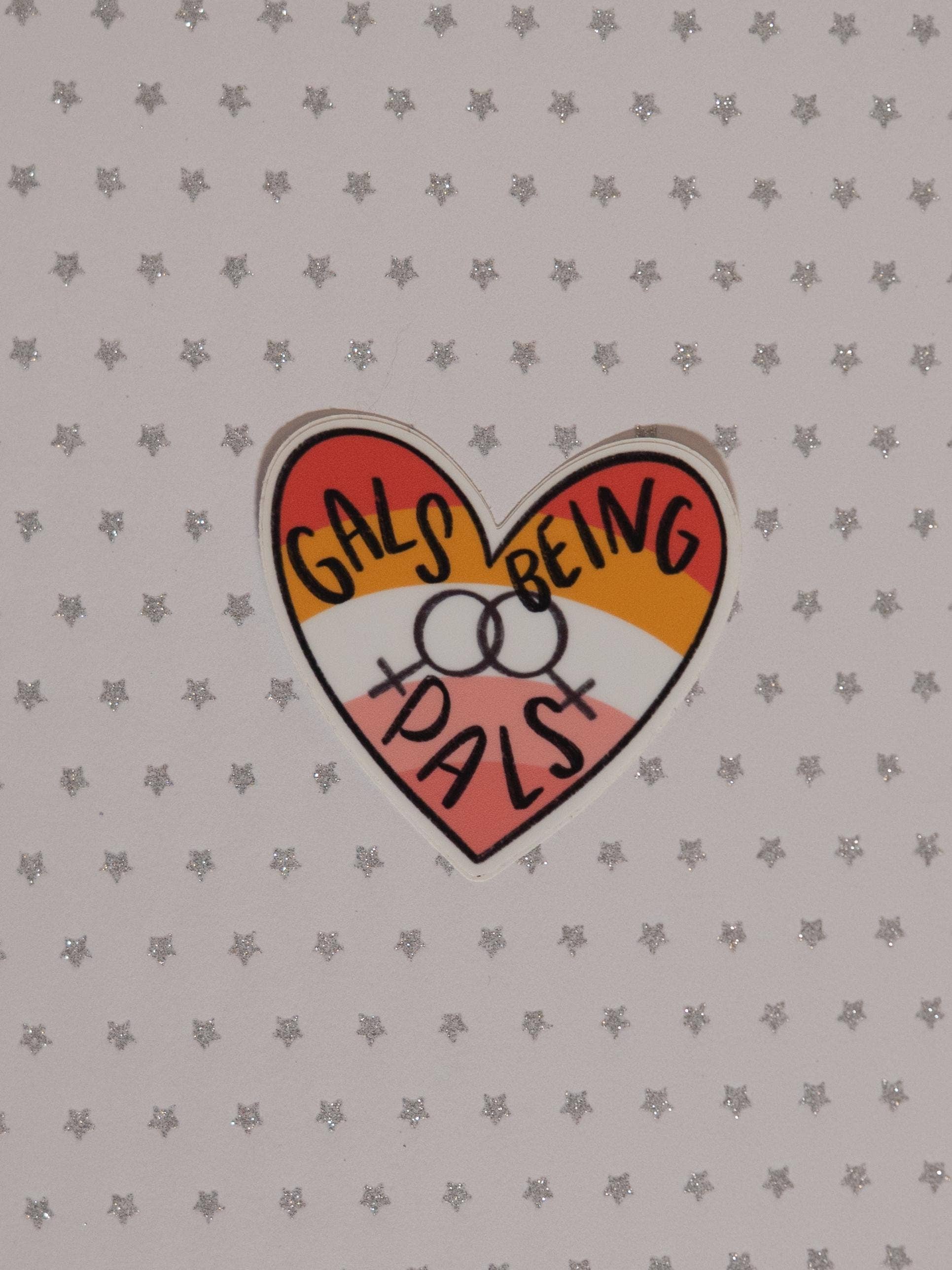 A heart-shaped sticker with the text 'GALS BEING PALS' in black handwritten-style lettering over red, orange, yellow, and pink sections, placed on a light gray star-patterned background.