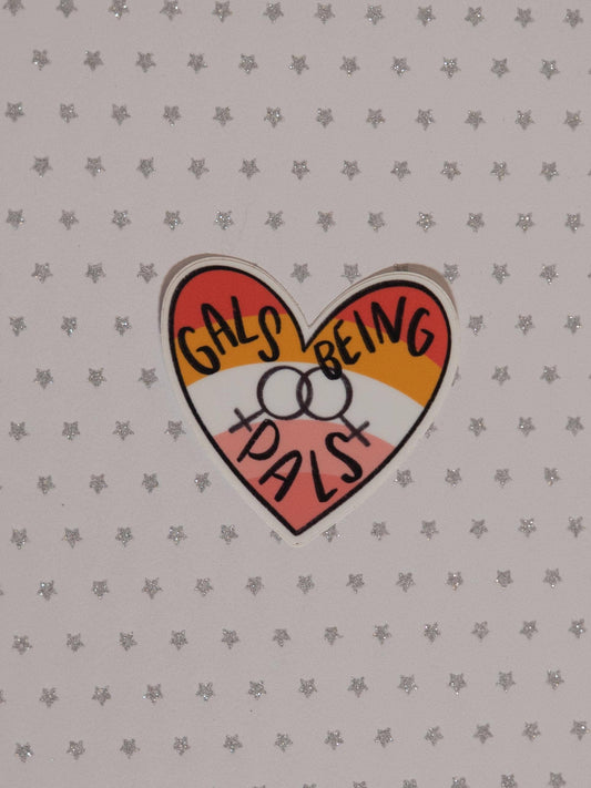 A heart-shaped sticker with the text 'GALS BEING PALS' in black handwritten-style lettering over red, orange, yellow, and pink sections, placed on a light gray star-patterned background.