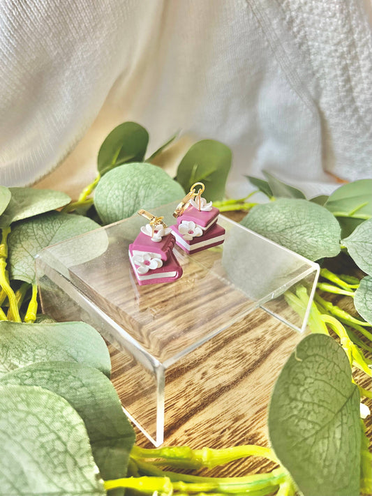 Pink book-shaped earrings on a wooden stand with green leaves in the background